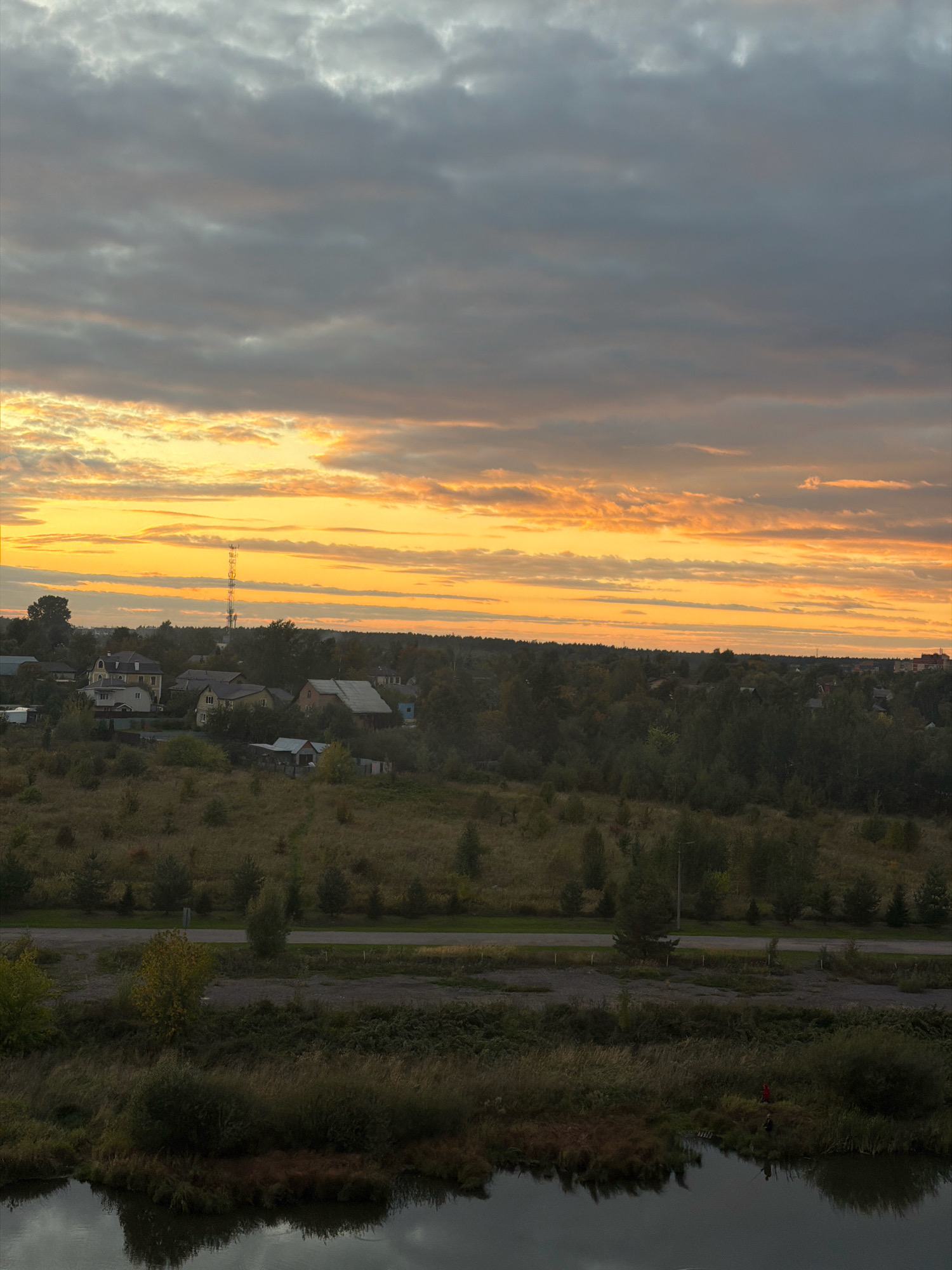 An aerial view captures the evening sky over Vniissok, Russia, during a sunset. The sky is filled with dramatic clouds, a mix of gray and orange hues indicating the setting sun. Below, a residential area with houses and trees is visible, surrounded by a dense forest. A telecommunications tower stands tall in the distance, a prominent feature against the colorful sky. The foreground shows a body of water reflecting the sky, with reeds and grass lining its banks. A small red object, possibly a person or a marker, is visible on the bank near the water. The overall scene depicts a tranquil rural landscape at dusk.