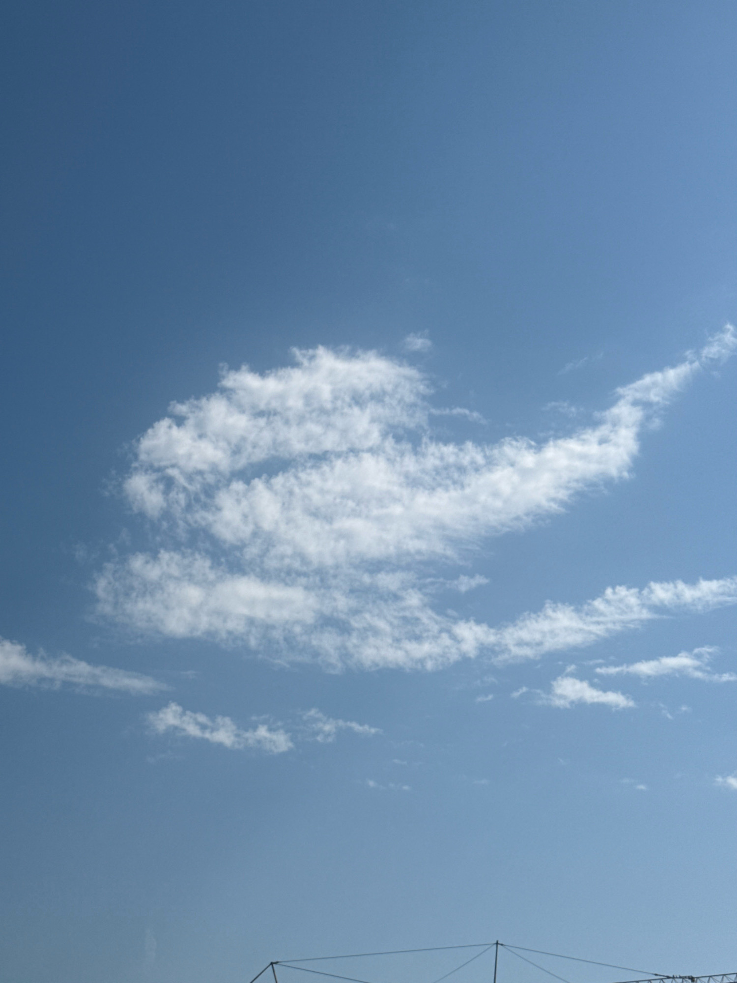 The image shows a clear blue sky with scattered white clouds. The clouds form a shape that resembles a bird in flight or a whale. In the bottom of the image, there is a glimpse of a dark structure that appears to be part of a crane or scaffolding, suggesting an outdoor urban environment. There are no people visible in the image. The location is identified as Boreti, Montenegro.