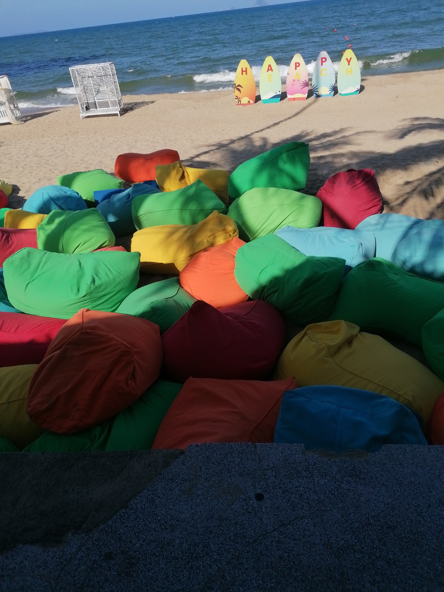 This photo, taken in Nha Trang Ward, Vietnam, shows a collection of colorful beanbag chairs on a beach. The chairs are predominantly green, blue, yellow, orange, and red. In the background, there is a sandy beach with ocean waves, a white beach chair, and five surfboard-shaped signs spelling out "HAPPY". The foreground is partially obscured by a textured gray surface, suggesting the photo was taken from an elevated position. Shadow patterns indicate the presence of trees or other structures casting shadows on the scene.