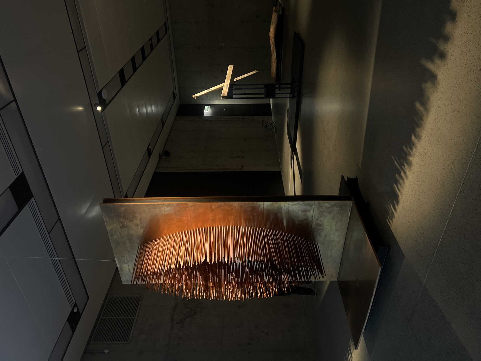 The image shows the interior of what appears to be the Garage Museum of Contemporary Art in Moscow, Russia. The perspective is angled upward, capturing the modern architectural elements and the art installations. The museum has a sleek and minimalist design, with visible ventilation and lighting systems.
A prominent artwork hangs from the ceiling: a rectangular structure supports numerous thin, vertical copper-colored rods suspended downward. Another installation, appearing to be made of wooden planks arranged in a cross-like formation, is visible in the background. The lighting is dramatic, with a combination of direct and ambient light creating areas of shadow and highlights on the textured walls and floor. No people are present in the frame. The composition draws the eye toward the interplay of geometric forms and the contrast between the raw materials and the refined architectural space.
