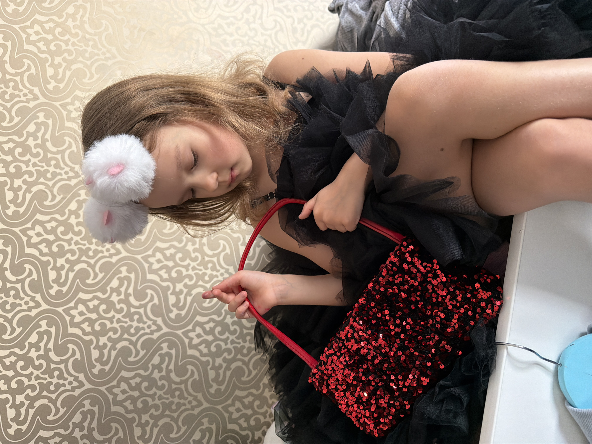 The image captures a young girl lying on her side, eyes closed, suggesting she is resting or asleep. She has light brown or blonde hair that falls around her face and shoulders. On her head, she wears a white headband adorned with two fluffy, spherical pom-poms that resemble bunny ears, each featuring a small pink patch in the center. A dark-colored, possibly black, choker or necklace is visible around her neck.
She is dressed in a black garment made of ruffled or layered tulle fabric, which appears to be a dress or a top. Her legs are curled up, and her bare skin is visible, surrounded by the black tulle. A small mole can be seen on her upper thigh.
The girl holds a vibrant red purse in her right hand, clutching its red strap. The purse is entirely covered in small, shimmering red sequins, giving it a festive and playful appearance.
The background primarily consists of a wall covered in light-colored (beige or cream) wallpaper featuring an ornate, flowing, and somewhat damask-like pattern in a slightly darker shade. To the right of the girl, a white surface is visible, possibly part of a bed frame or furniture. A light blue, circular object, perhaps a pair of headphones or a small speaker, with a silver metal hook or clip is also partially visible on this white surface.
The overall impression is one of peaceful rest, perhaps after a period of imaginative play, given her dress-up attire and accessories. The lighting appears soft, indicating an indoor setting. There is no visible text in the image.