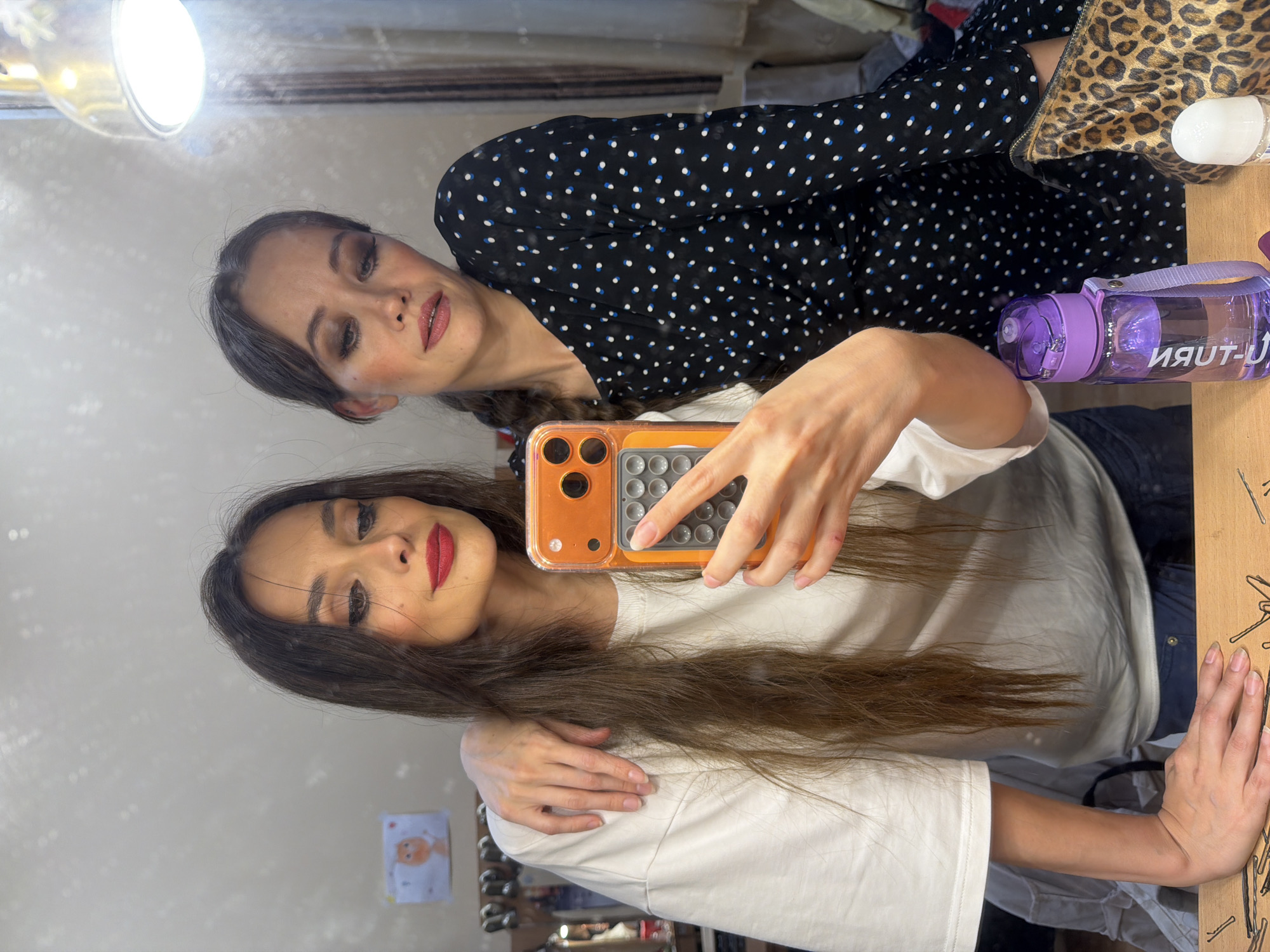 Two young women are taking a selfie in a room that appears to be a dressing room or backstage area. One woman, on the left, has long dark hair and is wearing a white t-shirt. The other woman, on the right, has her hair in a braid and is wearing a black dress with white and blue polka dots. They are both looking at the phone, which is being held by the woman on the left. In the background, there is a mirror, a bright light, and some shelves with various items. There is also a leopard print bag and a purple water bottle on a wooden surface in the foreground. The overall scene suggests a casual, behind-the-scenes moment, possibly before or after a performance or event. The location of Ufa, Russia, is not evident from the visual cues in the image.
