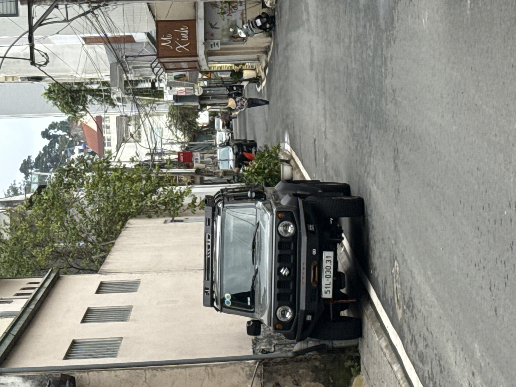 This image captures a vibrant street scene, likely in Da Lat, Vietnam, characterized by a narrow paved road, surrounding buildings, and abundant utility wires overhead. The overall impression is one of everyday life in a bustling urban or semi-urban environment.
In the foreground, prominently parked on the right side of the street, is a dark-colored Suzuki Jimny SUV, equipped with a roof rack and rugged tires. Its license plate, "51L-030.31," is clearly visible. The vehicle faces away from the viewer.
Further down the street, a person wearing a traditional Vietnamese conical hat (nón lá) and a patterned shirt walks away from the camera. Other figures and parked motorbikes are discernable in the distance. On the right side of the street, a brown wooden sign above a storefront reads "Mị Xinh" in script, suggesting a business. Below it, a gray scooter is parked.
To the left, a light-colored building with multiple rectangular ventilation openings is visible. Lush green foliage from trees or bushes adds a touch of nature amidst the concrete structures. The sky appears bright but somewhat hazy or overcast, indicating daytime. The jumble of overhead electrical or communication wires is a common feature in many Vietnamese cities. The scene depicts a typical, quiet moment of daily activity on a residential or commercial street.