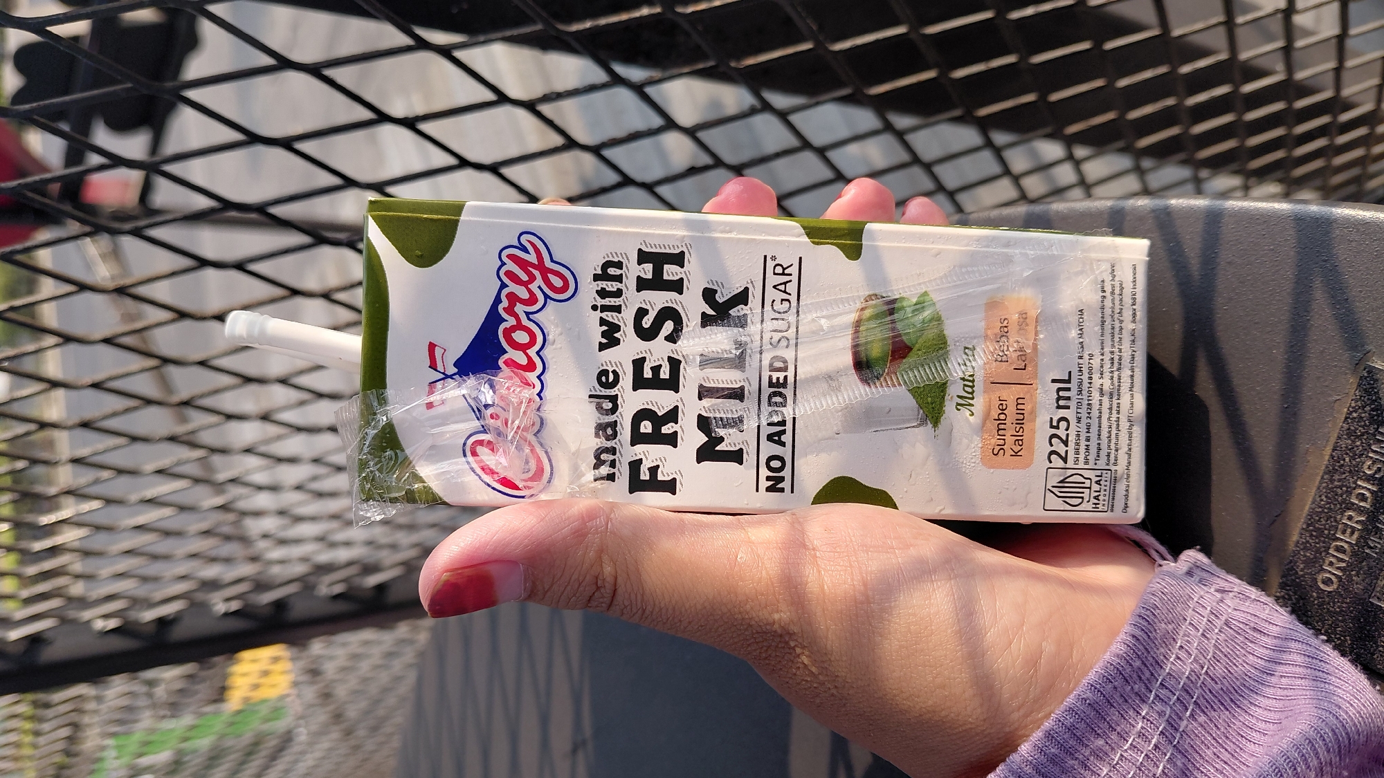 A person's hand, with painted red fingernails, holds a carton of "Good Day" milk. The carton is green and white and features text stating "made with FRESH MILK" and "NO ADDED SUGAR". Below this, it indicates the flavor is "Matcha", and that it is a "Sumber Kalsium" (Source of Calcium) and "Bebas Laktosa" (Lactose-Free). The carton also displays "225 mL" and a Halal certification. The carton is partially wrapped in clear plastic. The background shows a black metal fence and a blurred outdoor environment, suggesting it is daytime. The scene is outdoors, likely in an urban or semi-urban setting, possibly in the city of Karanglewas, Indonesia, given the Halal certification and Indonesian language text on the product.