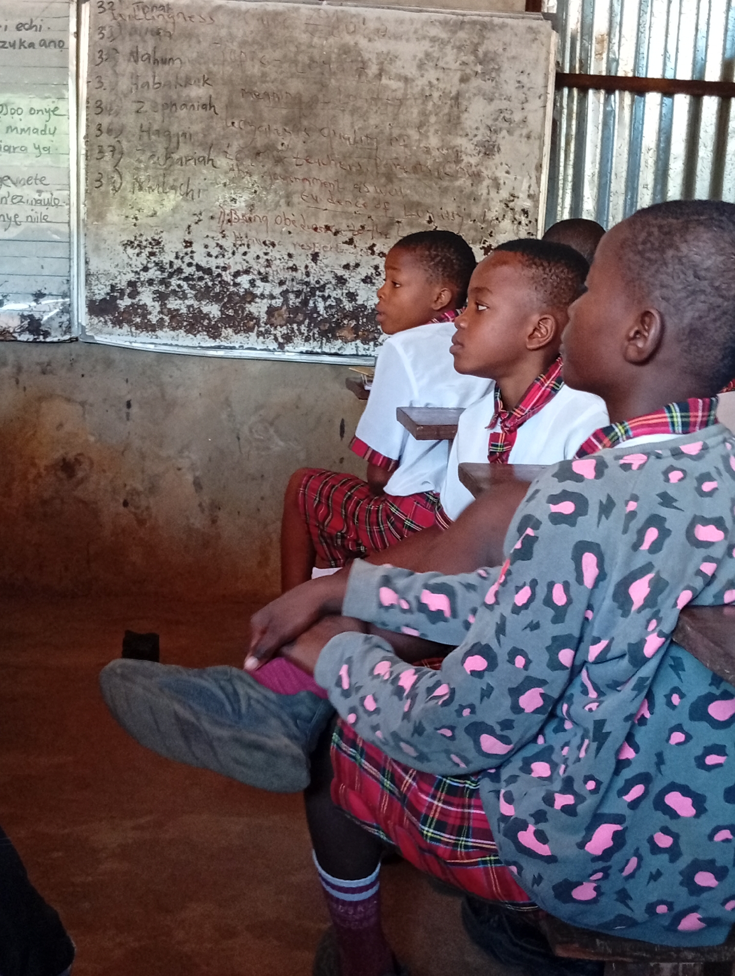 The image shows three Black children sitting in a classroom. They wear school uniforms with white shirts, plaid neckties, and plaid skirts or shorts. The child in the foreground has a gray sweatshirt with pink leopard spots. The children are looking to the left. Behind them is a dirty chalkboard with writing in chalk. The classroom has concrete walls, and a corrugated metal roof is visible. The location is likely in a rural area.