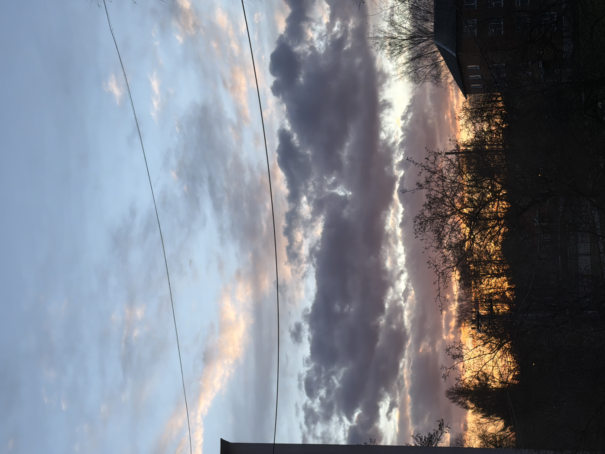 The image depicts a sky during a sunset in what appears to be an urban setting. A power line runs diagonally across the sky. The sky is filled with clouds that transition in color from a light blue and pink on the left side to darker, grey hues in the center, and a vibrant orange-pink on the right side, indicating the presence of the setting sun. The silhouette of trees and a building are visible in the right portion of the image, while a rooftop is seen on the lower part of the image, obscuring part of the scene. Based on the location information provided, it is likely a view of the sky over Reshetnikovo, Russia, with the obscured trees and rooftops being part of the urban landscape. No people or actions are visible in the frame.