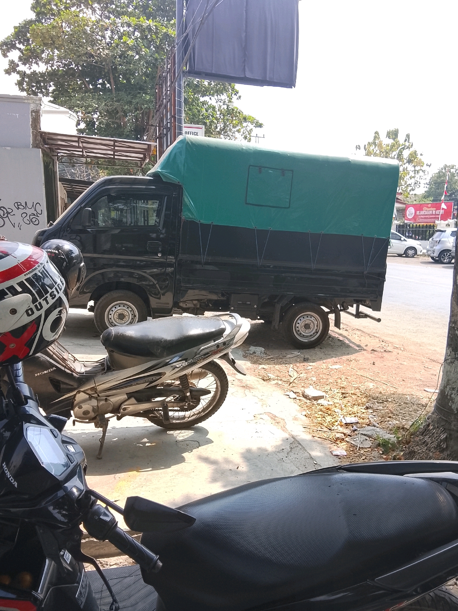In Slawi, Indonesia, a black pickup truck with a green tarp covering its bed is parked next to a motorcycle. The truck is on a dirt and paved road, with trees and buildings in the background. The motorcycle is silver and black, with a red and white helmet resting on its handlebars. The scene appears to be outdoors on a sunny day.