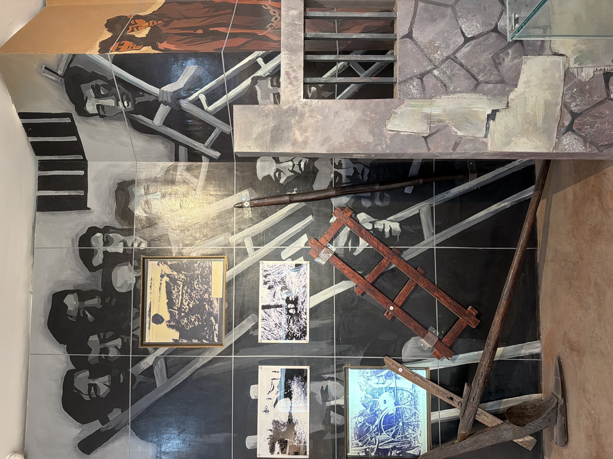 The image depicts an interior scene likely within a museum or historical exhibit in Thuận Hóa Ward, Vietnam. The walls are covered in murals painted in shades of gray, black, and white, featuring stylized depictions of individuals, some appearing to be prisoners or laborers, in various poses. Several framed black and white photographs are affixed to the murals, some of which appear to depict historical events or scenes related to work and hardship. In the foreground, on the floor, lie several old agricultural tools, including a plow and a wooden ladder-like structure. The overall atmosphere suggests a historical commemoration of difficult times, potentially related to labor or imprisonment.