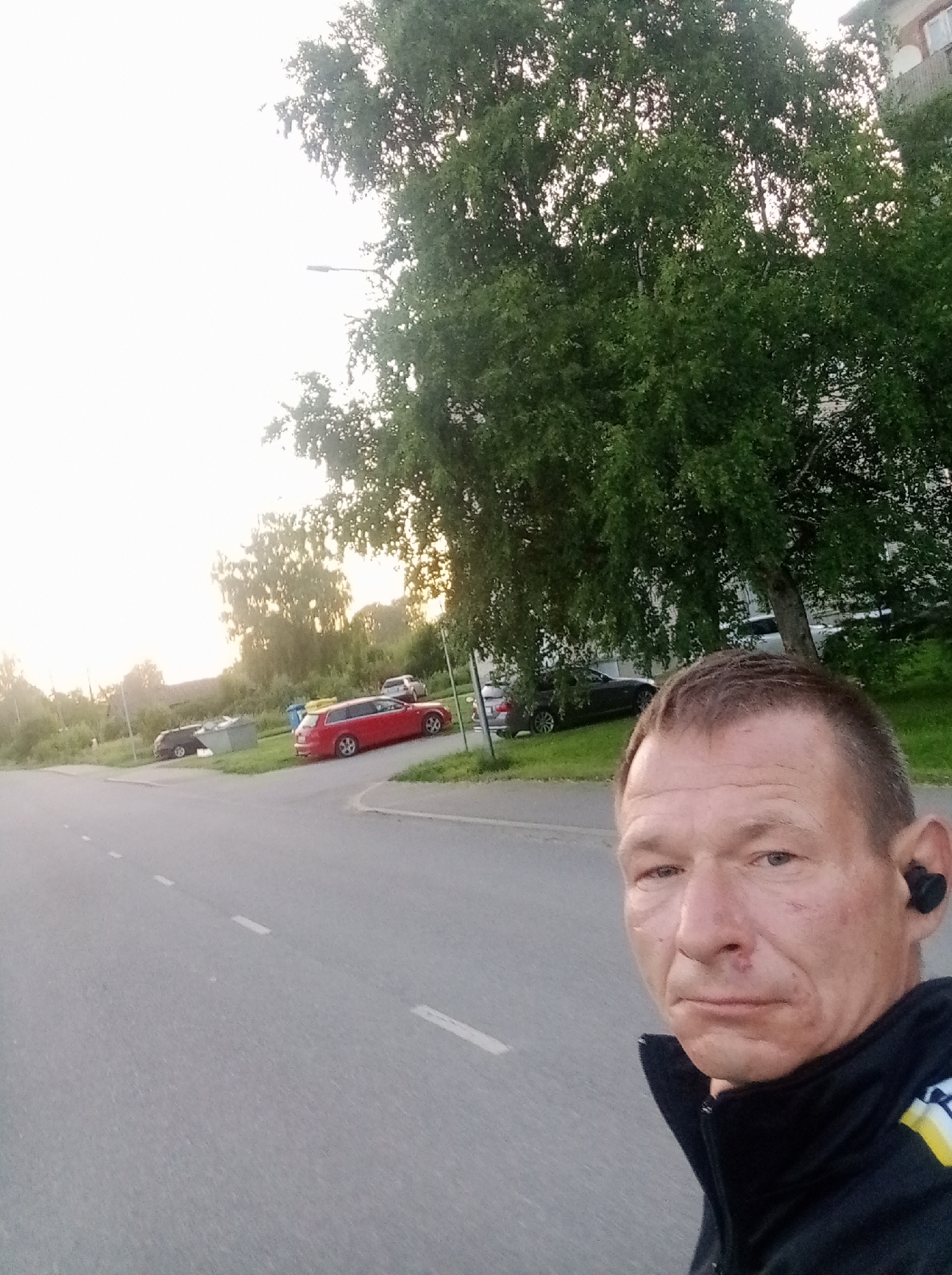 A selfie of a man with short brown hair and light skin is taken outdoors. He is wearing a black jacket with yellow and white accents on the sleeve. He has black earbuds in his ears and what appears to be a small cut or scab on his nose. He is looking directly at the camera with a neutral expression.

The background shows a paved road with white dashed lane markings. To the left of the road, there is a grassy area with several parked cars, including a red station wagon and a dark sedan. In the distance, there are trees and what looks like a building. A street light is visible in the upper left portion of the image. The sky is bright and hazy, suggesting it might be dawn or dusk, or a cloudy day.

The location is identified as Tapa, Estonia. The scene depicts a street with cars and trees, indicating a residential or semi-urban area. The man appears to be on a walk or run, given the outdoor setting and athletic attire.