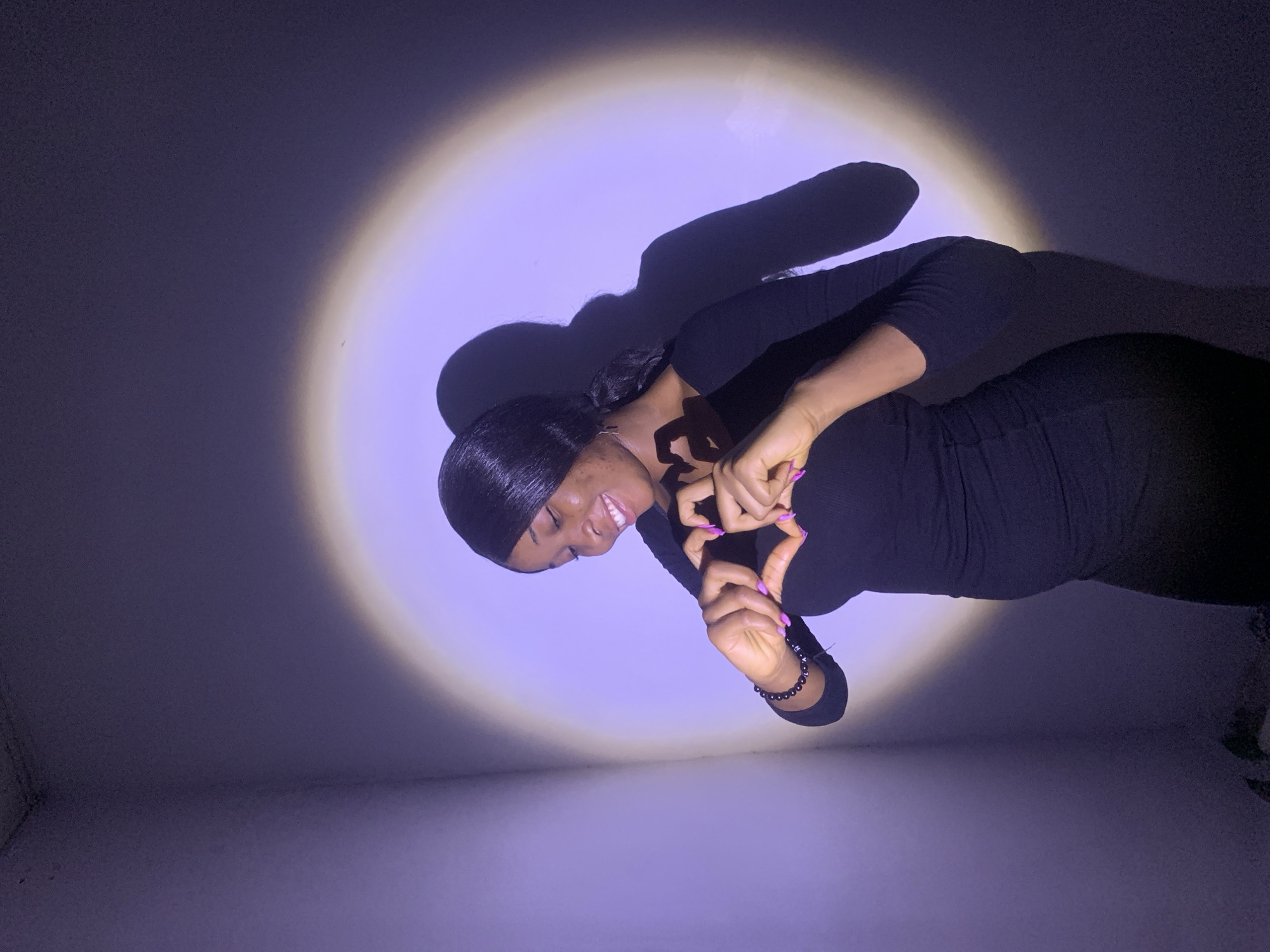 A young woman with dark hair pulled back is posing indoors. She is wearing a long-sleeved black dress and making a heart shape with her hands. She's smiling and facing slightly to the left. A light shines on her, casting her shadow on the wall behind her. The location is likely indoors, but there is no visible information to establish the city of Jos, Nigeria.