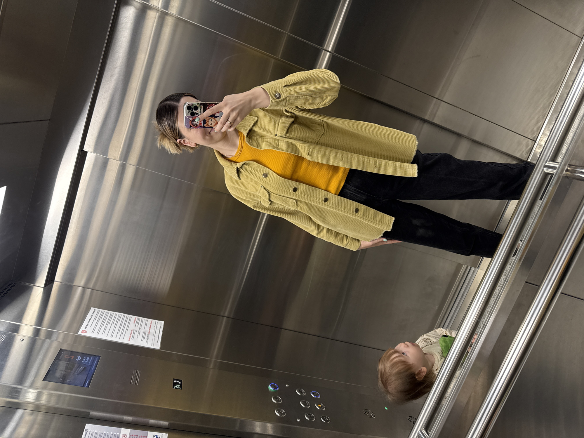 The image, rotated counter-clockwise, captures a selfie taken inside a modern elevator with a young adult and the reflection of a child. The elevator interior features brushed stainless steel walls, a control panel with several illuminated blue buttons, and a metal handrail on the right.
The primary subject is a young adult, likely female, holding a smartphone with a colorful, possibly anime-themed, case to take the picture. She has short, light brown hair styled with a side part. She is wearing a bright yellow ribbed long-sleeve top under a light olive green/mustard yellow corduroy jacket, paired with black pants. A ring is visible on her left hand.
In the lower right portion of the image, the reflection of a young child, possibly a toddler, is visible in the metallic wall. The child has light brown hair and is wearing a light-colored, patterned long-sleeved top, appearing to hold a green object.
Above the elevator buttons, a small digital screen displays "SON" and some partially readable text that includes what looks like a floor number (e.g., "этаж 02"). Two white information sheets are affixed to the elevator wall. The upper sheet features a red heart icon and the Russian text "АКТУАЛЬНАЯ ИНФОРМАЦИЯ И АКЦИИ" (Current Information and Promotions). The smaller text on this and a partially visible lower sheet is also in Russian. The setting is an indoor, artificially lit environment, consistent with an elevator in a building in Moscow, Russia.