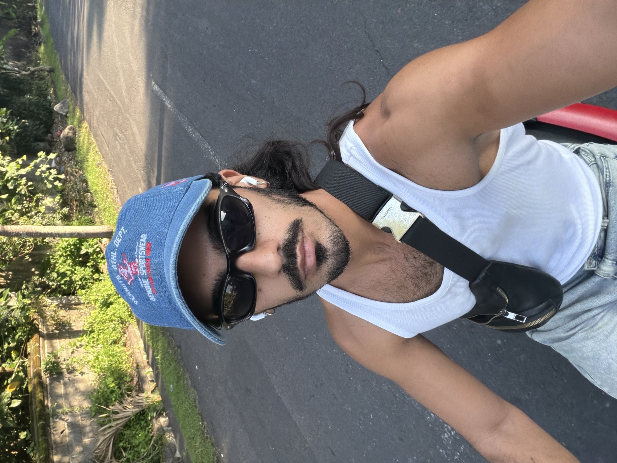 A person is captured in a sunlit outdoor selfie, likely while stationary or riding on an asphalt road in North Kuta, Indonesia. The photo is taken from a slightly elevated perspective, looking down.
The subject is a person with dark, long hair, styled back, and wearing a white ribbed tank top, light-wash jeans, and a blue denim baseball cap. They have a neatly trimmed beard and mustache, dark sunglasses, and white earbuds. A black fanny pack or crossbody bag with a metallic zipper and a prominent silver buckle is worn across the chest. The buckle clearly displays the text "CALZETTI." The denim cap features red embroidered text that reads, "ATHL. DEPT." and "PEANUTS GENUINE SPORTSWEAR."
The setting appears to be a sunny day, with clear skies and strong shadows visible on the asphalt road. To the left of the road, there's lush green foliage and tropical vegetation, consistent with the reported location context. A red object, possibly part of a vehicle or seat, is visible on the far right side of the frame. The person maintains a neutral expression, characteristic of a casual selfie.
