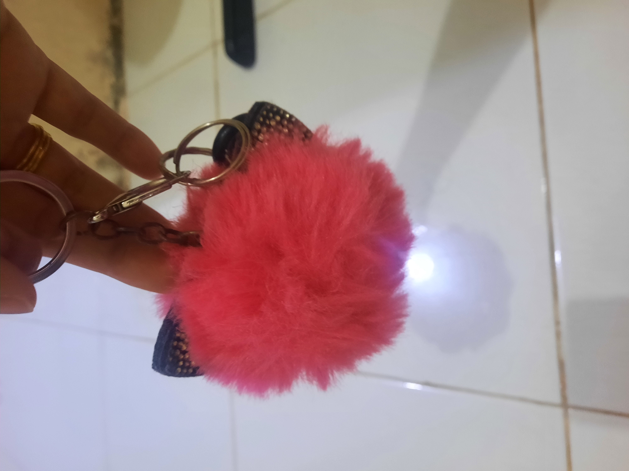 A close-up shot shows a person's hand holding a fluffy, bright pink keychain pom-pom with a sparkly black accent. The keychain is attached to a silver ring and chain. The background appears to be a tiled floor with thin grout lines, suggesting an indoor setting. The lighting is soft, with a bright reflection visible on the floor near the pom-pom, hinting at a light source. The overall impression is casual and personal, likely a snapshot of a personal accessory. The presence of a gold ring on the person's finger suggests the individual might be female. The location context is Lhokseumawe, Indonesia, but there are no specific visual cues in the image to confirm this. No text is visible in the image.