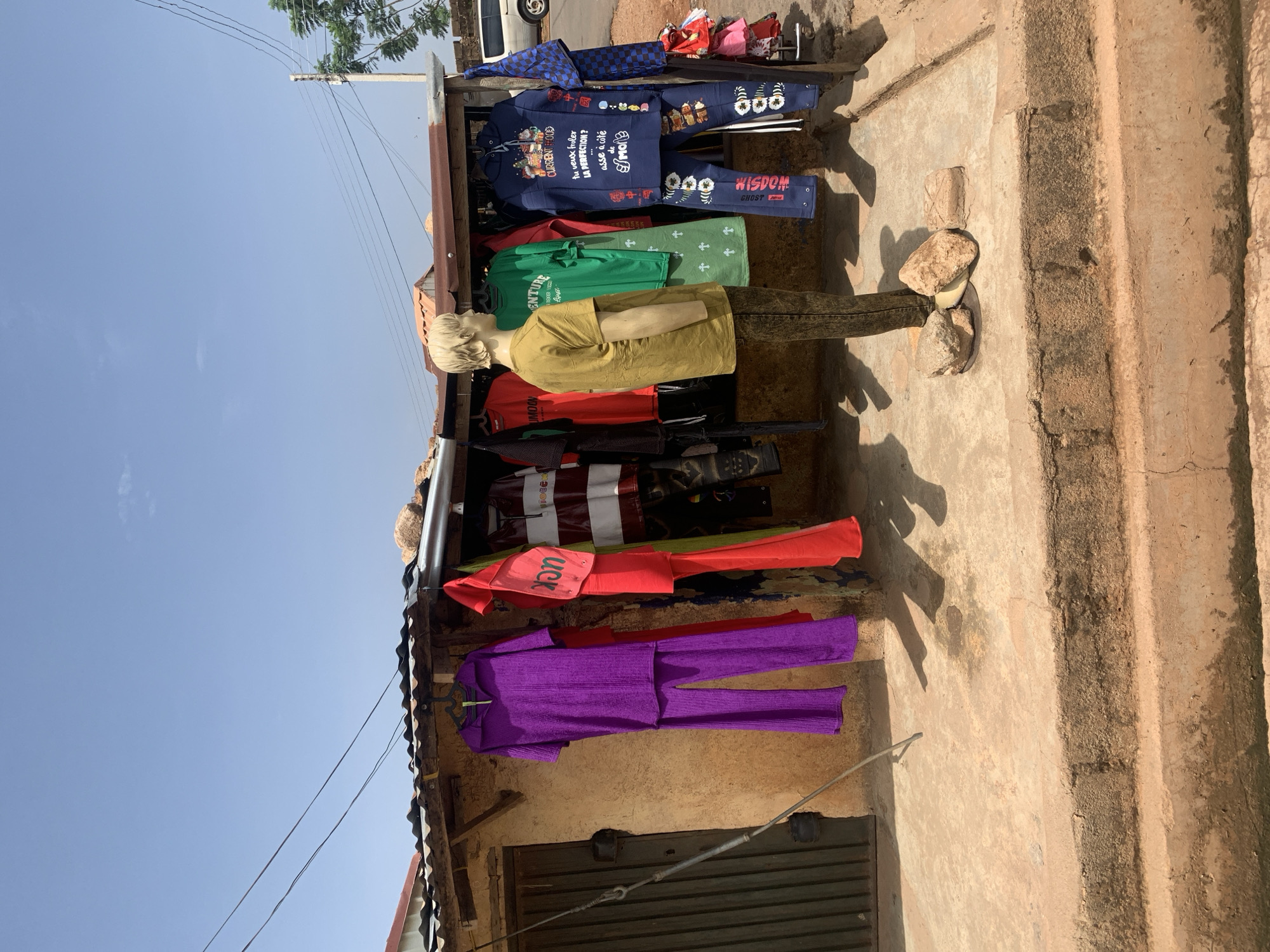 The image depicts a roadside clothing stall in Gura Top, Nigeria. Garments of various colors and styles, including purple, red, green, and blue outfits, are displayed on hangers. A mannequin stands in front of the stall, wearing a yellow shirt. The stall appears to be constructed with a corrugated metal roof and is located adjacent to a building with a weathered facade. The ground is partially paved and includes a small set of steps. Power lines stretch across the top portion of the frame, visible against the blue sky.