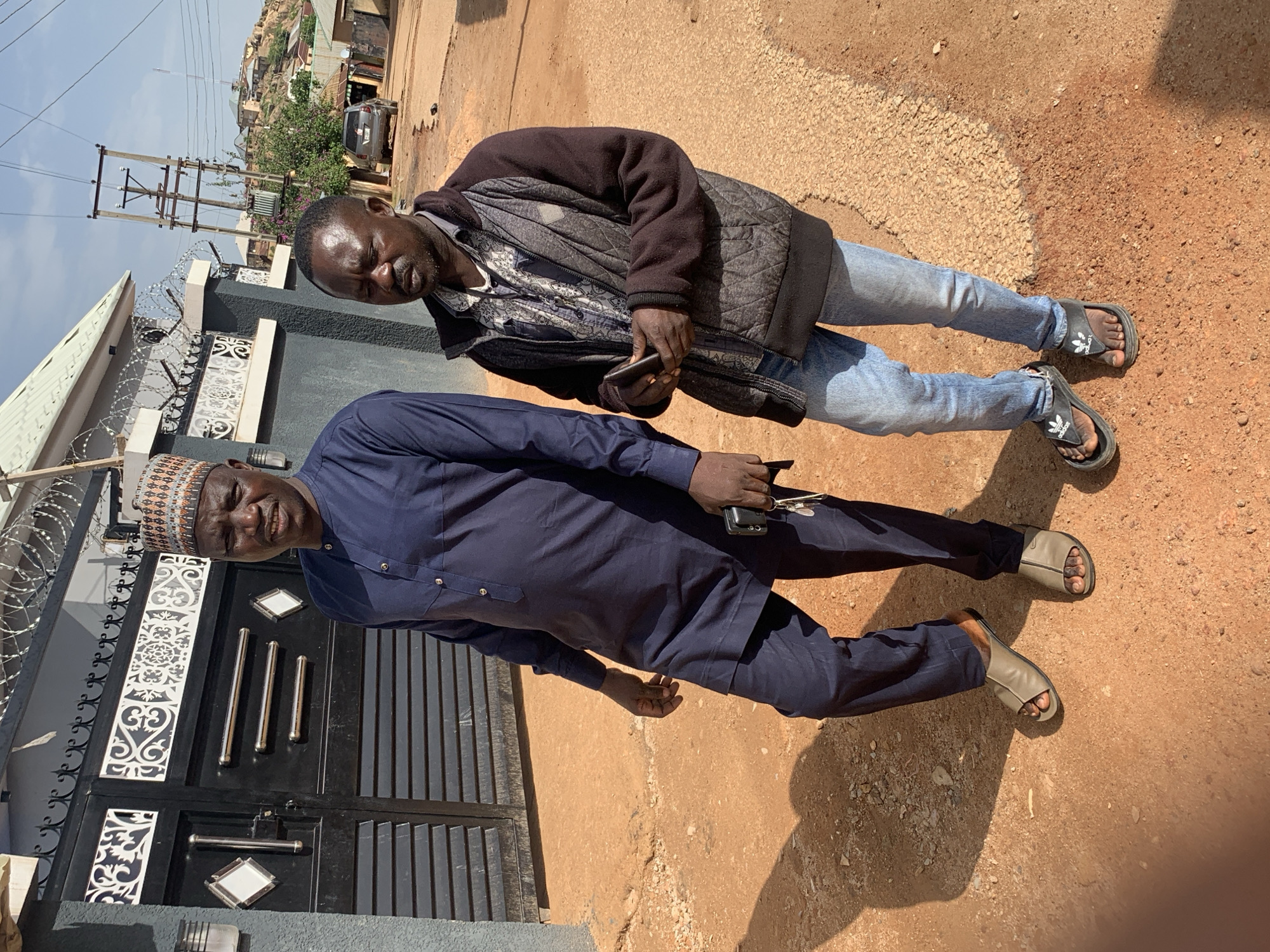 Two men are standing outdoors in what appears to be a residential area in Jos, Nigeria. The man in the foreground is wearing a dark blue outfit consisting of a long-sleeved shirt with a collar and matching pants, along with a patterned cap. He is looking towards the camera with a slight smile. Behind him and to his left, another man is visible. He is wearing a dark quilted jacket over a patterned shirt and light blue jeans. He is holding a dark-colored mobile phone in his right hand.
Both men are wearing open-toed sandals. The ground is a mix of dirt and gravel, with some scattered stones. In the background, there is a black metal gate with decorative white inserts and vertical metal bars. To the left of the gate, there is a grey wall with what looks like barbed wire on top. Further in the background, there are buildings, vegetation, and utility poles with electrical wires. The scene suggests a daytime setting, likely under clear skies, given the bright lighting and shadows. The overall mood seems casual and perhaps friendly, with the man in the foreground smiling. No visible text is present in the image.