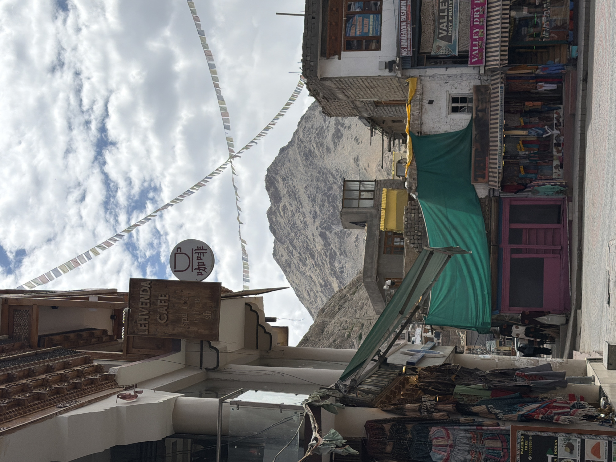 This is a street view in Leh, India, with a focus on a building facade on the left, shops in the mid-ground, and a mountain in the background. On the left, an ornately carved wooden overhang extends from a building. Below this, a wooden sign reads "LEHVENDA CAFE" with smaller text beneath that is partially obscured. To the right of the sign, a circular emblem with a coffee cup icon and Chinese characters is visible. Prayer flags are strung across the sky in diagonal lines.
In the mid-ground, various shops line the street. One shop features a vibrant green tarp covering part of its facade. A storefront with a pink exterior is visible below this tarp. Other shops display clothing and textiles hanging outside. Signs for "VALLEY DRY FRUITS" and "M/S HIMALAYAN PASHMINA" are prominent. Two individuals are walking on the street, one in a white shirt and the other in darker clothing. The ground is paved with cobblestones.
The background is dominated by a large, rugged mountain, its slopes covered in scree and rock. The sky above is cloudy with patches of blue visible. The overall impression is a lively market street in a mountainous region.