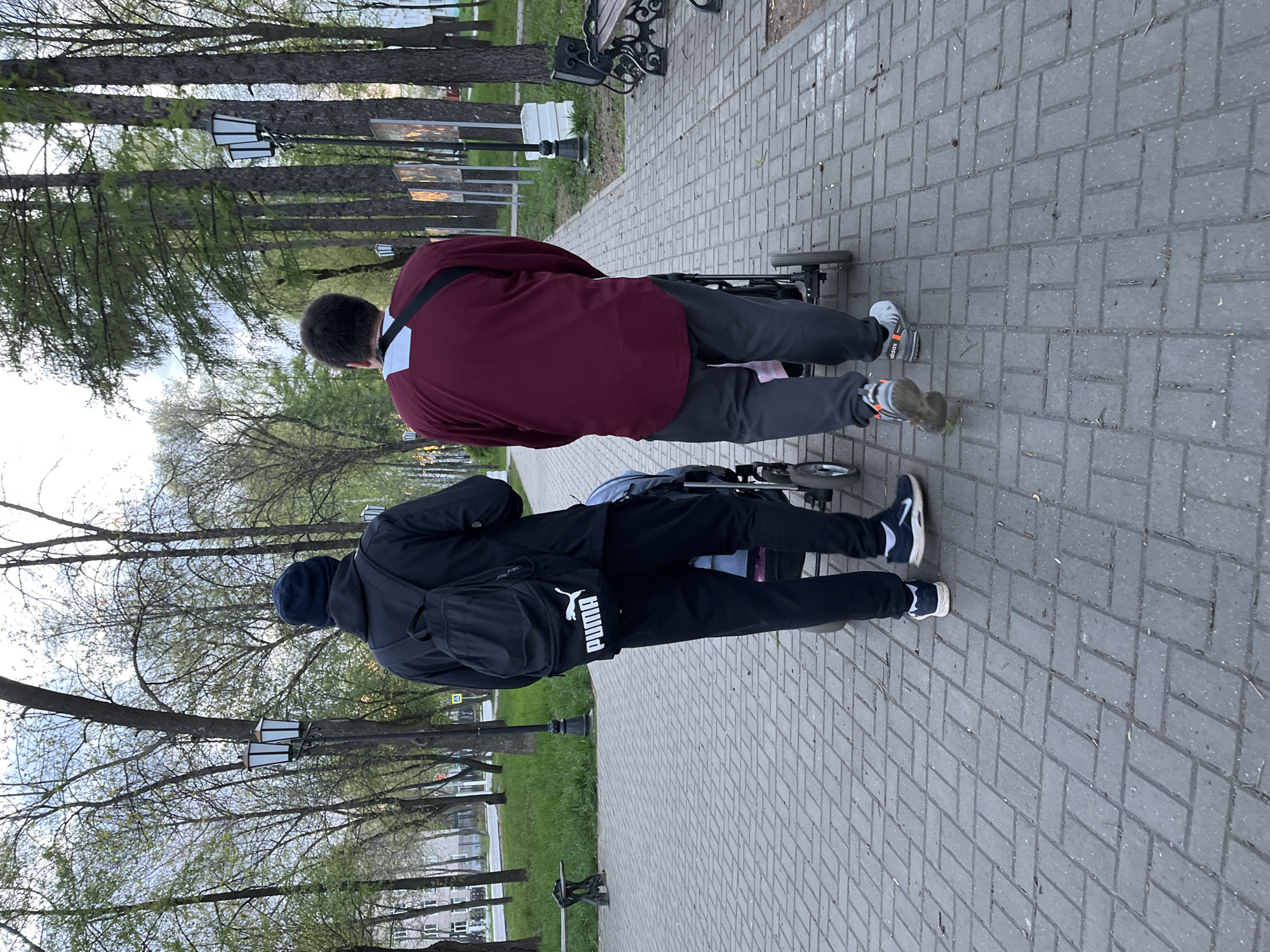 Two people walk on a paved path in Chaikovsky, Russia, pushing a stroller. The path, made of grey rectangular pavers, is lined with green grass and trees. One person wears a dark blue jacket, black Puma track pants, and a dark blue cap, carrying a black backpack. The other person wears a maroon shirt and grey pants. White street lamps are visible along the path and in the background. Benches are also seen in the background.