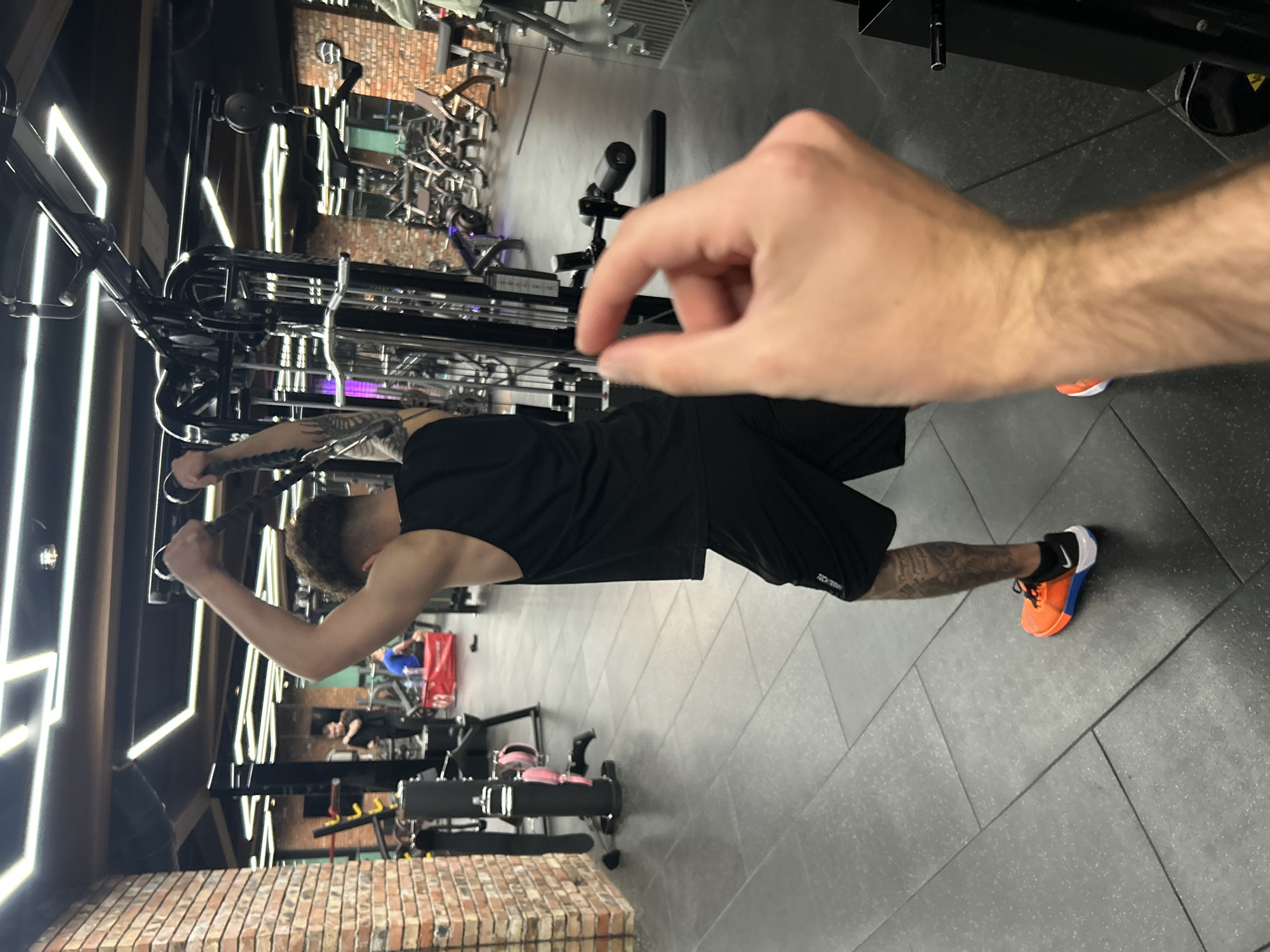 A man in a black tank top and black shorts is performing a lat pulldown exercise in a gym. He is pulling down on a rope attached to a cable machine. His shoes are brightly colored: orange, blue, black, and white. The gym has grey floors and mirrored walls. In the background, other gym equipment and people can be seen. A second person's hand and arm are visible, extended toward the camera and slightly blurred, and appearing to hold something between two fingers. The location is likely in Sofia, Bulgaria.