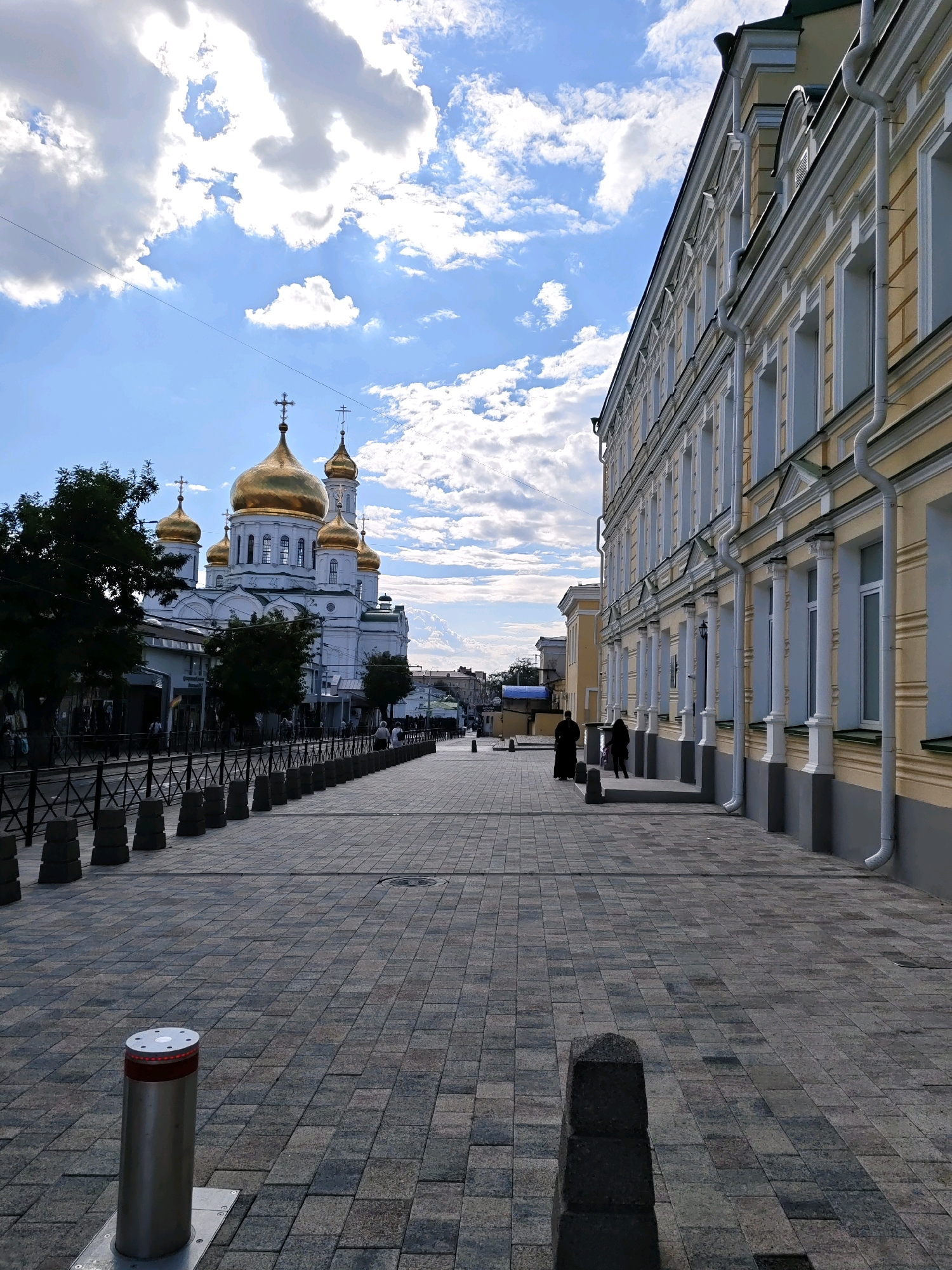 The image captures a view of a paved pedestrian walkway in Rostov-on-Don, Russia, looking towards a grand white church with golden domes. The church, likely the Nativity Cathedral, is prominent on the left side of the frame, its multiple gilded domes reflecting the daylight. Adjacent to the walkway, on the right, is a long, light-yellow building with numerous windows and architectural detailing, characterized by white vertical downspouts.
The walkway is paved with rectangular stone tiles in varying shades of gray and brown. A row of dark, metal bollards with some visible red lighting elements is positioned along the left edge of the walkway, bordering an area with trees and storefronts that are partially obscured by the trees. Further down the walkway, a few people are visible, two of whom are standing near the entrance of the yellow building, appearing to converse. The sky above is a bright blue, with scattered, fluffy white clouds. The overall impression is of an urban scene on a bright day, highlighting the architecture of the church and the adjacent building.