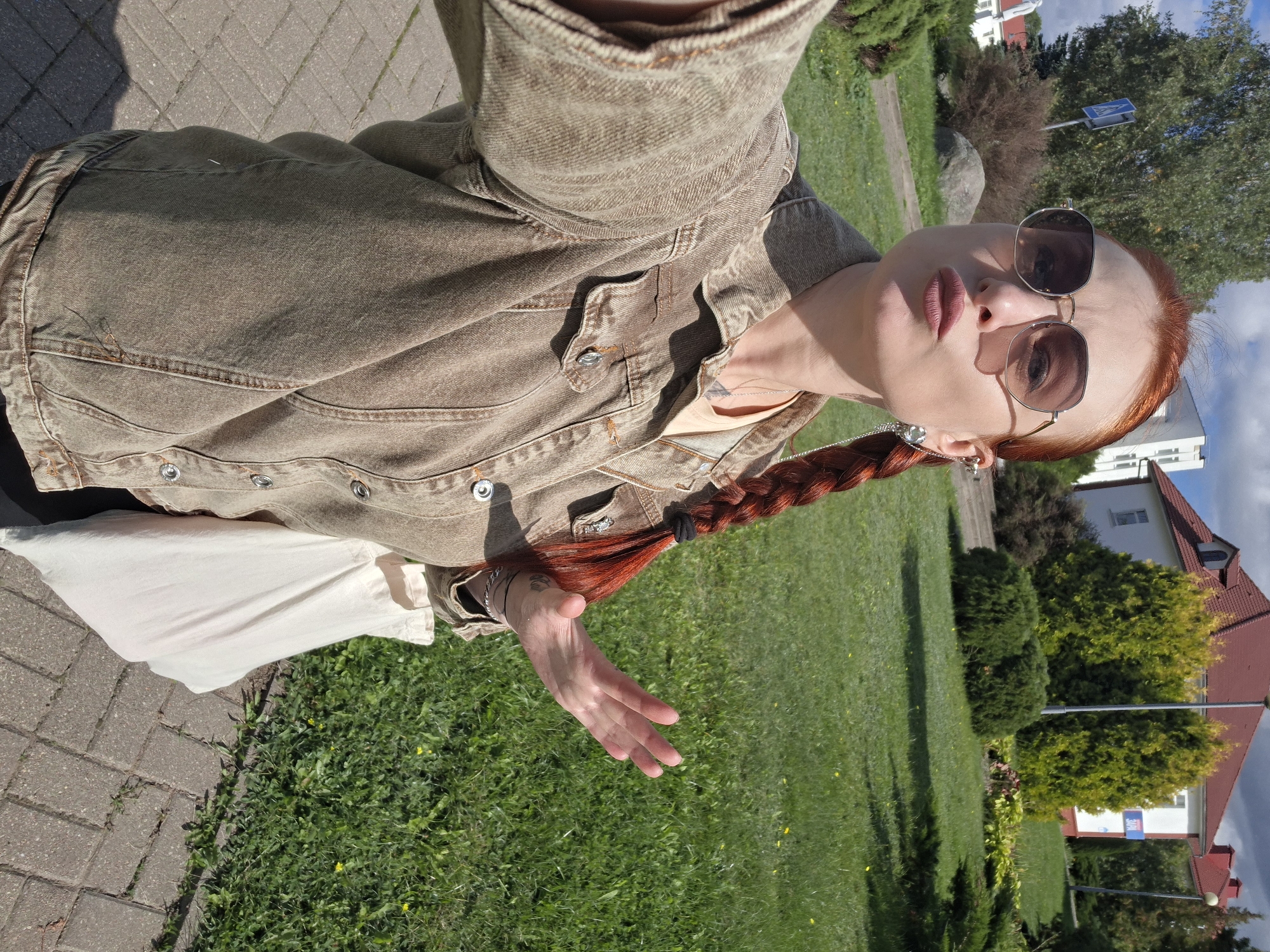 A woman with red hair, styled in a braid, is taking a selfie outdoors. She is wearing sunglasses, a denim jacket, and a light-colored t-shirt. She is holding a white tote bag in her left hand and has her right hand raised, palm facing outward. The background shows a grassy area with trees and buildings, suggesting an urban or suburban environment. The presence of a blue and white sign with an arrow and a white statue in the distance indicates the location is likely in or around the town of Myadzyel, Belarus, possibly near a park or public space. The day appears to be sunny with some clouds in the sky.