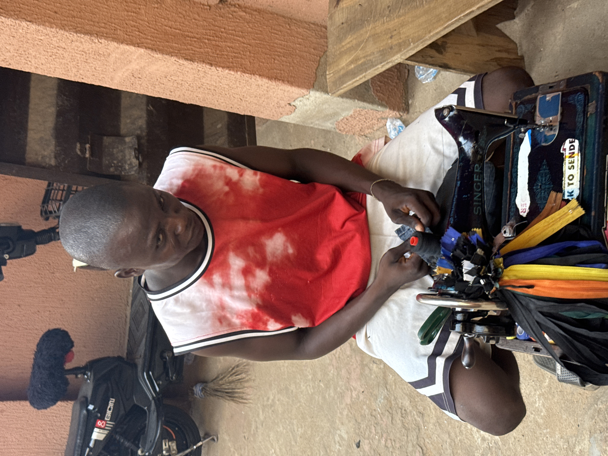 The image captures a man, likely a tailor or seamster, intently operating a Singer sewing machine in what appears to be an outdoor or semi-sheltered workspace. He is seated on a low surface, wearing a red and white tie-dye-patterned sleeveless shirt and light-colored pants with dark vertical stripes. His attention is focused downwards on the fabric he is working on, with a small object, possibly a spool of thread or part of the garment, held in his left hand.
The robust, dark-colored sewing machine is a prominent object, with "SINGER" visible on its side. It is adorned with a collection of colorful zippers in various hues including black, yellow, orange, blue, and green, suggesting he specializes in zipper repairs or related items. A sticker on the machine displays the partial text "K TO SENDE".
To the man's left, a portion of a black motorcycle or scooter is visible, bearing the brand name "GICIN". A fluffy black object, possibly a microphone windsock or a cleaning duster, is attached to its handlebar. The ground around him is an earthy, light brown, with scattered plastic water bottles and a broom nearby. The background consists of a textured, reddish-brown wall or pillar.
The scene depicts a diligent artisan engaged in his craft during what appears to be daytime, indicated by the clear, natural lighting. The setting conveys a practical, functional workshop environment, consistent with street-side commerce in Onitsha, Nigeria.