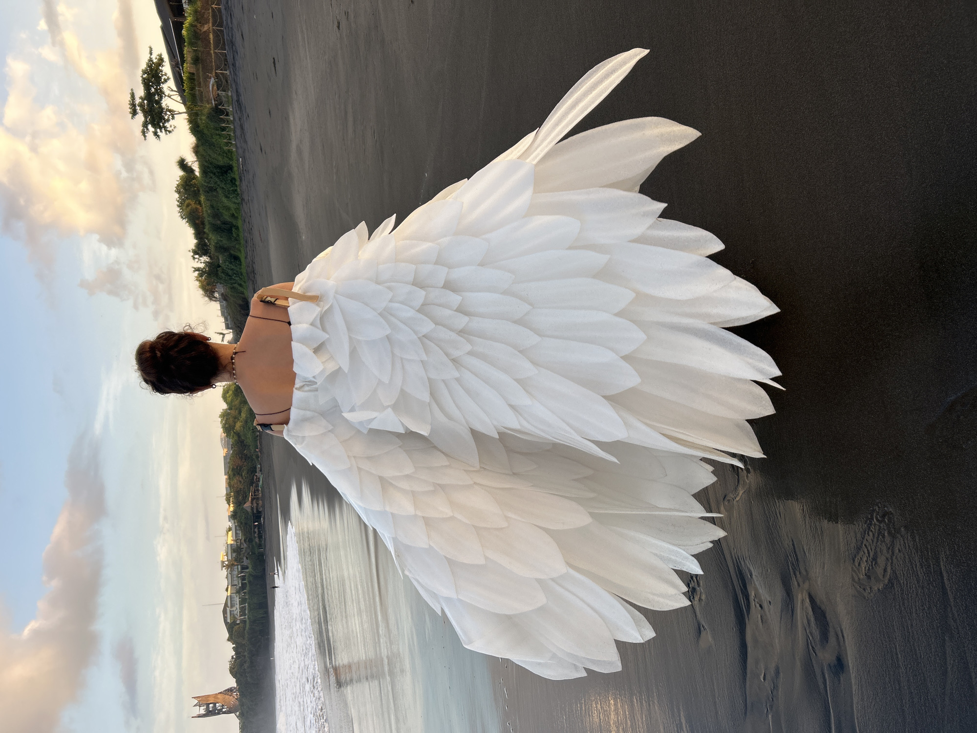 An image captures a person from behind, standing on a dark sandy beach during either sunrise or sunset. The individual is adorned with a large, elaborate garment resembling voluminous white wings or a feathered skirt, constructed from numerous layered, petal-like fabric pieces that extend widely behind them. Their dark hair is styled in an updo, and a necklace or strap is visible around their neck and shoulders. A small, fan-like wooden object is discernible near their back, possibly integrated into the white attire.
The scene features a tranquil beach with dark, possibly volcanic sand, showing subtle textures and ripples. To the right, gentle waves from a body of water, likely the ocean, meet the shore. In the distant background, a coastline with lush green vegetation, including trees and bushes, along with several buildings, stretches along the horizon. The sky is illuminated with soft, warm tones of yellow, orange, and blue, characteristic of the "golden hour," casting a serene and ethereal glow over the landscape. The overall composition suggests a moment of contemplation or a dramatic, artful pose. No text is visible in the image.