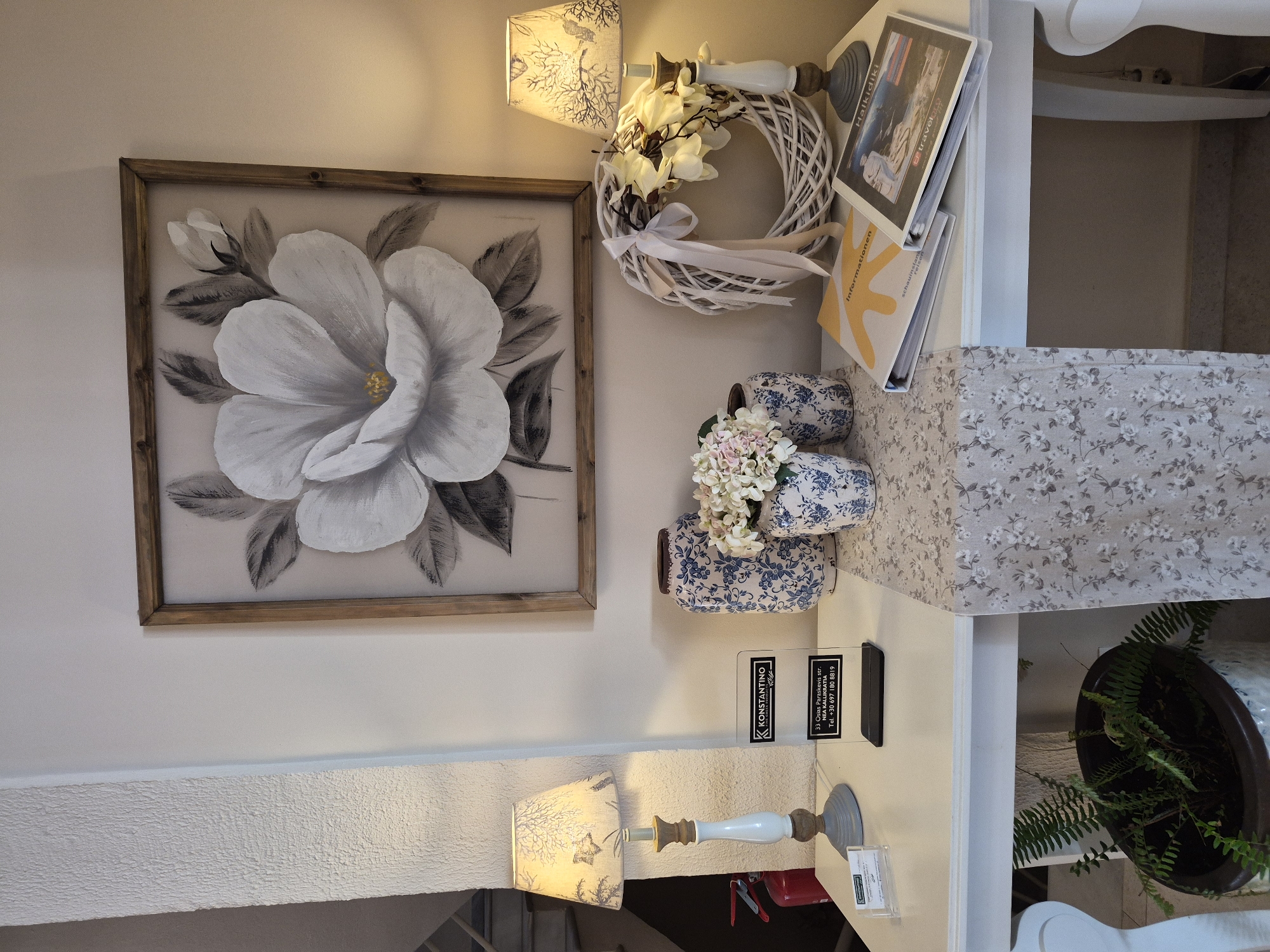 The image depicts a well-lit interior scene, likely a reception area or shop display, located in Néa Kallikrátia, Greece. A white table serves as the central display surface. On the left, a table lamp with a patterned shade sits atop a white and gray base. To its right, a clear acrylic sign displays business information, including the name "KONSTANTINO" and an address: "33 Osias Paraskevis str. NEA KALLIKRATIA." Two blue and white patterned ceramic vases are positioned on the table; one holds a bouquet of light pink and white flowers, while the other is empty. A white wicker wreath adorned with white fabric flowers and ribbons is placed to the right of the vases. A gray and white floral runner stretches across the table. Two open binders are on the right side of the table; one has a yellow cover with the word "Informationen" and the other features an image of people on a beach. A potted fern is visible in the lower left corner. The background features a large framed artwork of a black and white floral painting hanging on a beige wall. To the left, a textured white column and a red fire extinguisher are partially visible. The overall ambiance is clean and decorative.
