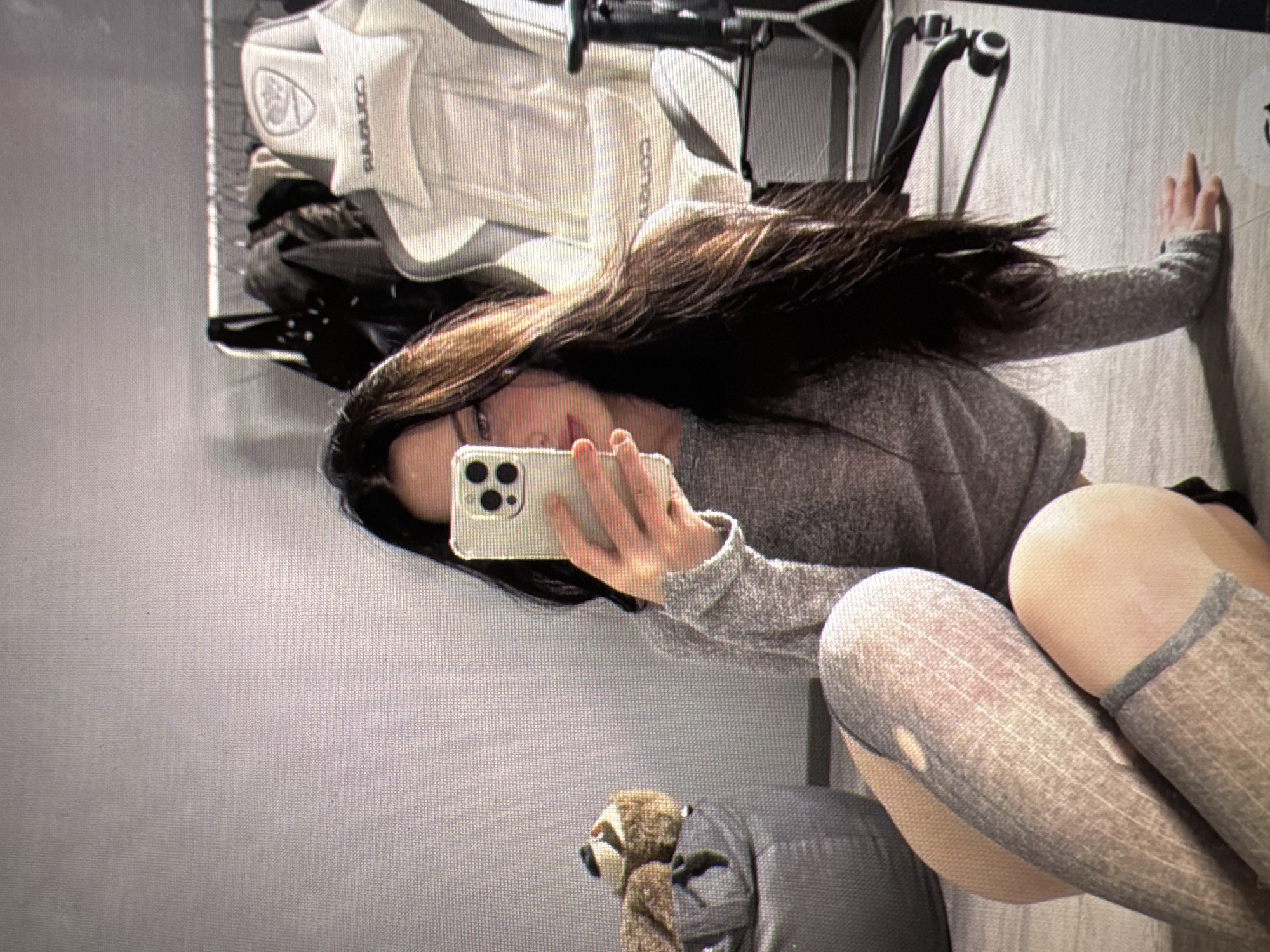A young woman with long, dark hair is taking a selfie with her phone in a room. She is wearing a grey, long-sleeved shirt and grey thigh-high stockings with a distressed pattern. Her legs are crossed as she sits on a wooden floor. To her left is a grey pouf with two stuffed animals on top. In the background, a white gaming chair with a black frame and wheels is visible, along with a rack of clothing. The overall impression is of a casual indoor setting, possibly a bedroom or living space. There is no discernible information to place the image in Castelo Branco, Portugal.