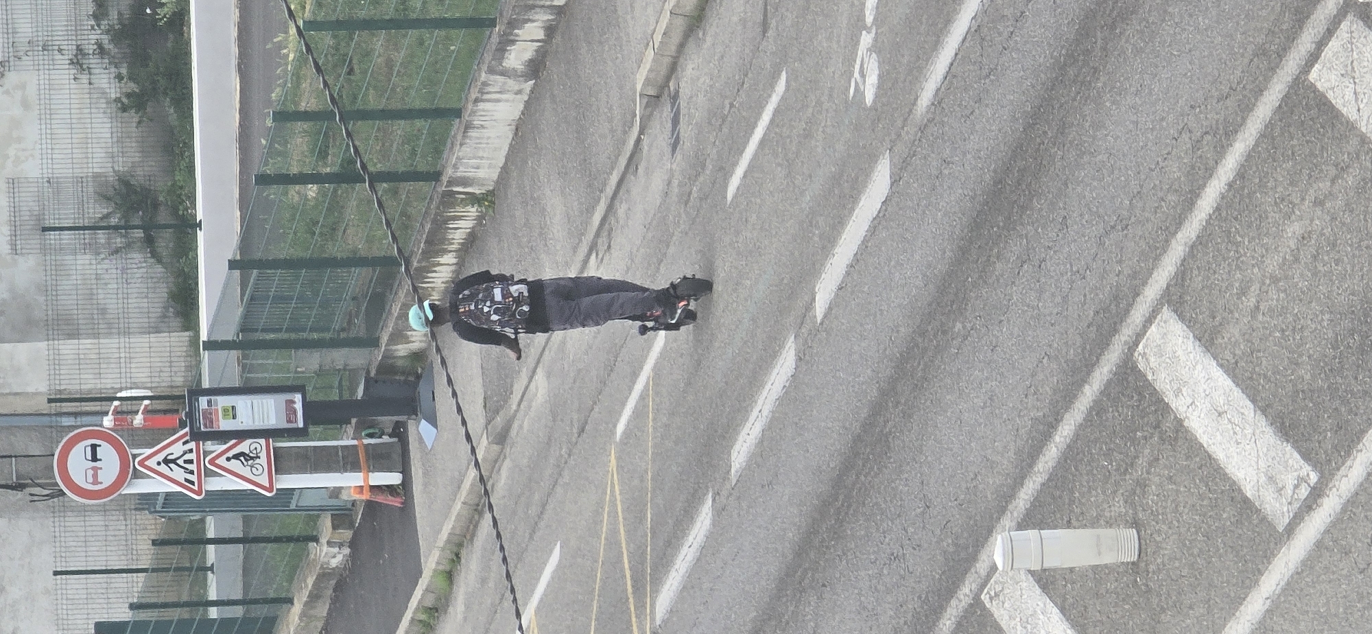 The image shows a person riding an electric scooter on a road. The person is wearing a light blue cap and a backpack. The scooter is black and appears to be moving forward. There are several road signs visible in the background, including a "no overtaking" sign, a pedestrian crossing sign, and a bicycle crossing sign. There is also a bus stop sign. The road is asphalt and has white lane markings. The environment appears to be urban, with a fence and buildings visible in the background. The lighting suggests it is daytime.