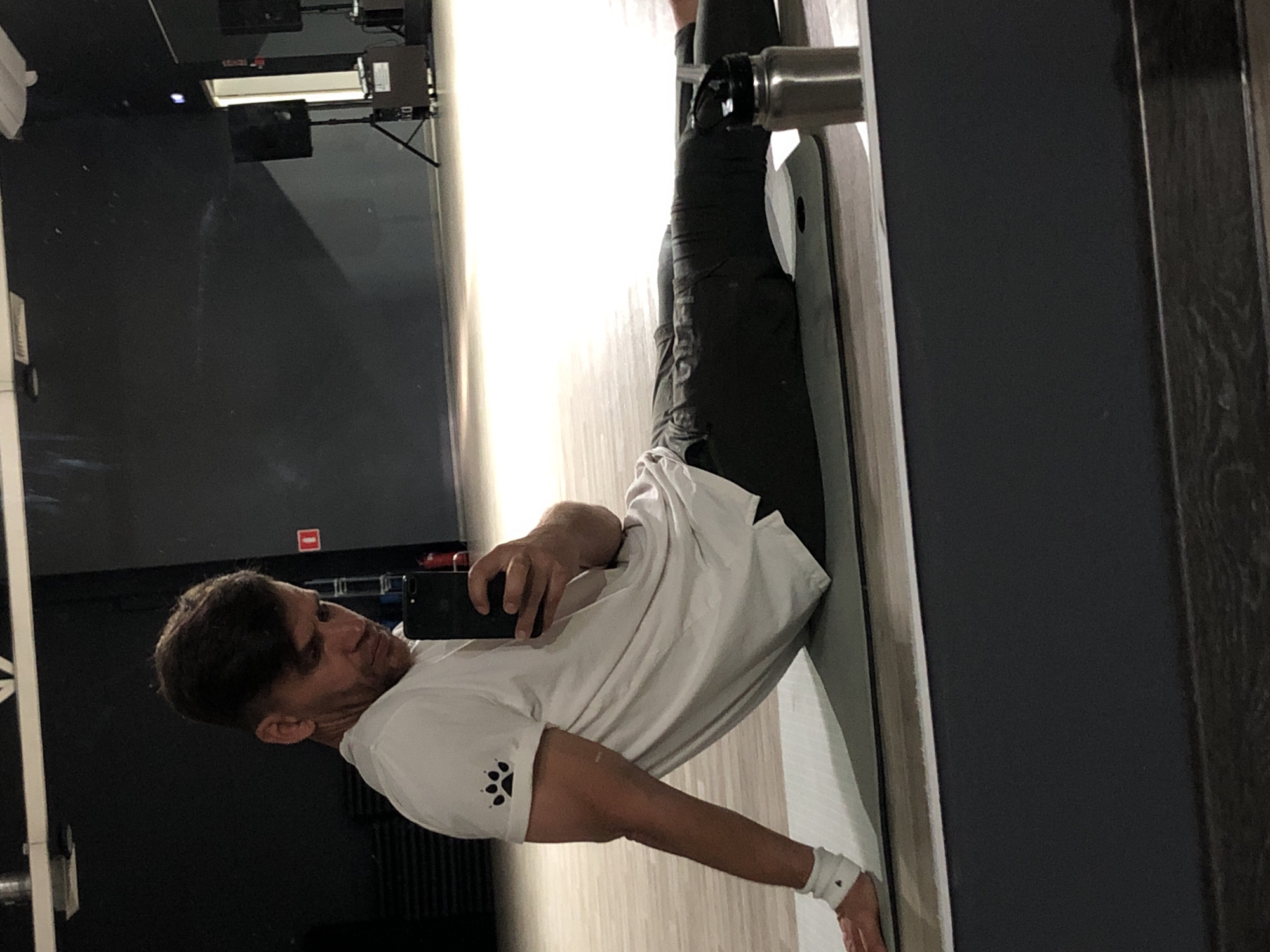 A man with short, dark hair and stubble is taking a selfie in a gym. He is wearing a white t-shirt with a black paw print logo on the left sleeve and dark grey pants. He is sitting on a grey yoga mat with his legs extended and his torso leaning back slightly, supported by his left arm. A white wristband is visible on his left wrist. A stainless steel water bottle with a black lid is positioned to his right. The gym has dark walls and wooden flooring. In the background, there are speakers on stands and some gym equipment. The scene is reflected in a large mirror, creating a sense of depth and showing the photographer's perspective. The lighting is dim, with some areas brightly illuminated. The image appears to be taken indoors, possibly in Saint Petersburg, Russia.