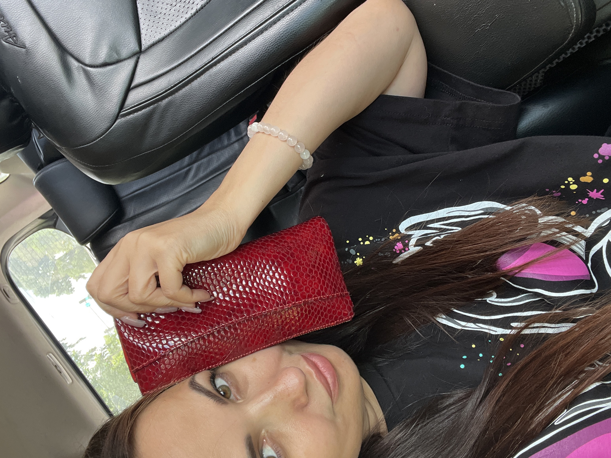 A woman with long, dark brown hair sits in the passenger seat of a car, holding a red, textured clutch to her cheek. She is wearing a black t-shirt with a white and pink graphic design. A beaded bracelet adorns her left wrist. The car's interior, upholstered in black leather, is visible behind her. The background suggests an outdoor environment, possibly with trees and a glimpse of a traffic light. The scene is brightly lit, likely by daylight.
