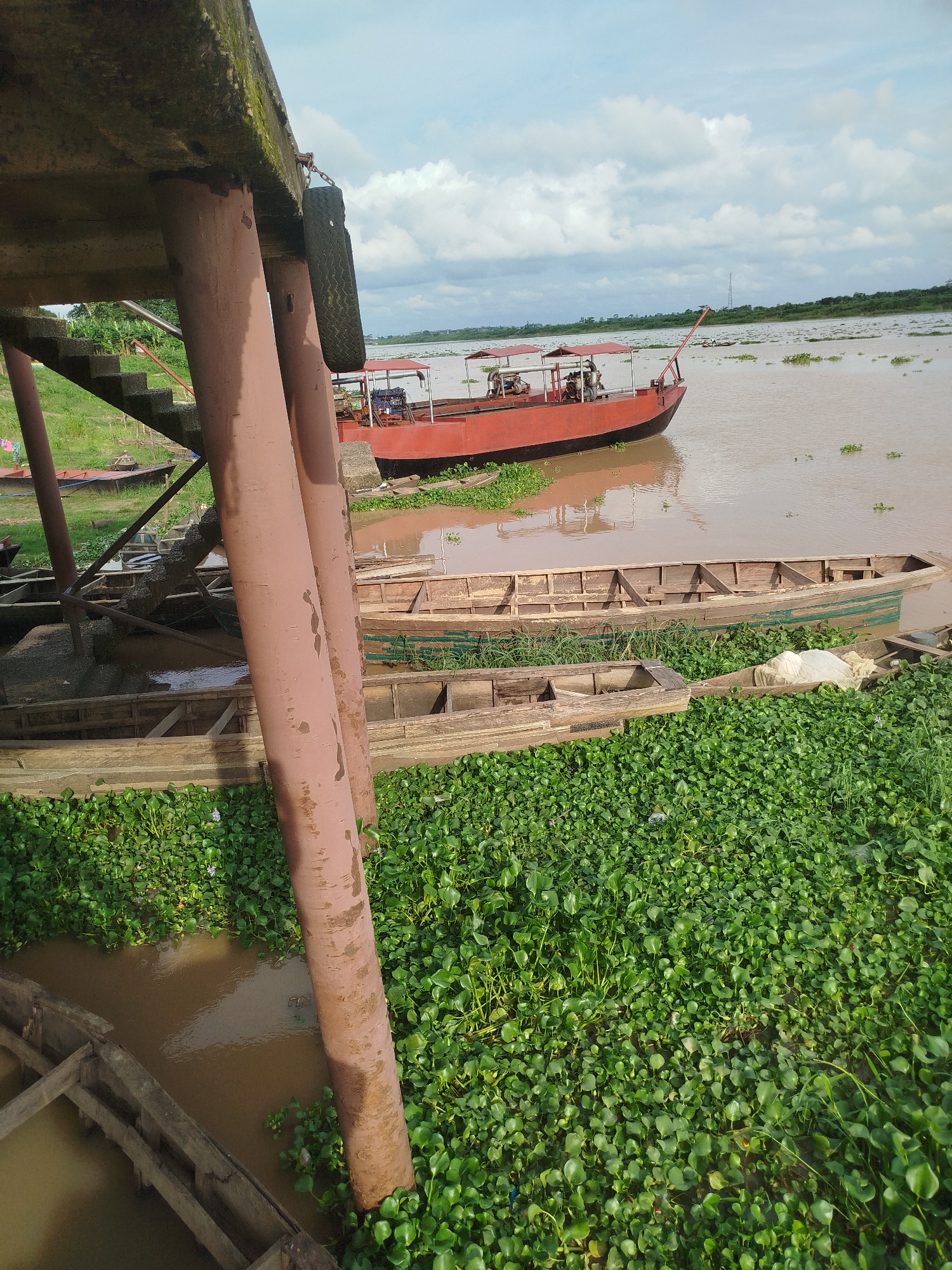 In Yenagoa, Nigeria, a murky river is lined with abundant green aquatic plants, creating a dense carpet on the water's surface. Several wooden canoes are partially submerged and rest near the bank, some appearing worn and weathered. A larger, red boat with a covered canopy is moored in the water further out. The scene is partially framed by the underside of a concrete structure supported by large, reddish-brown metal pillars. A tire is hanging from a chain on one of these pillars, possibly as a fender. In the background, the river widens, with a distant treeline and a hazy sky with scattered clouds.
