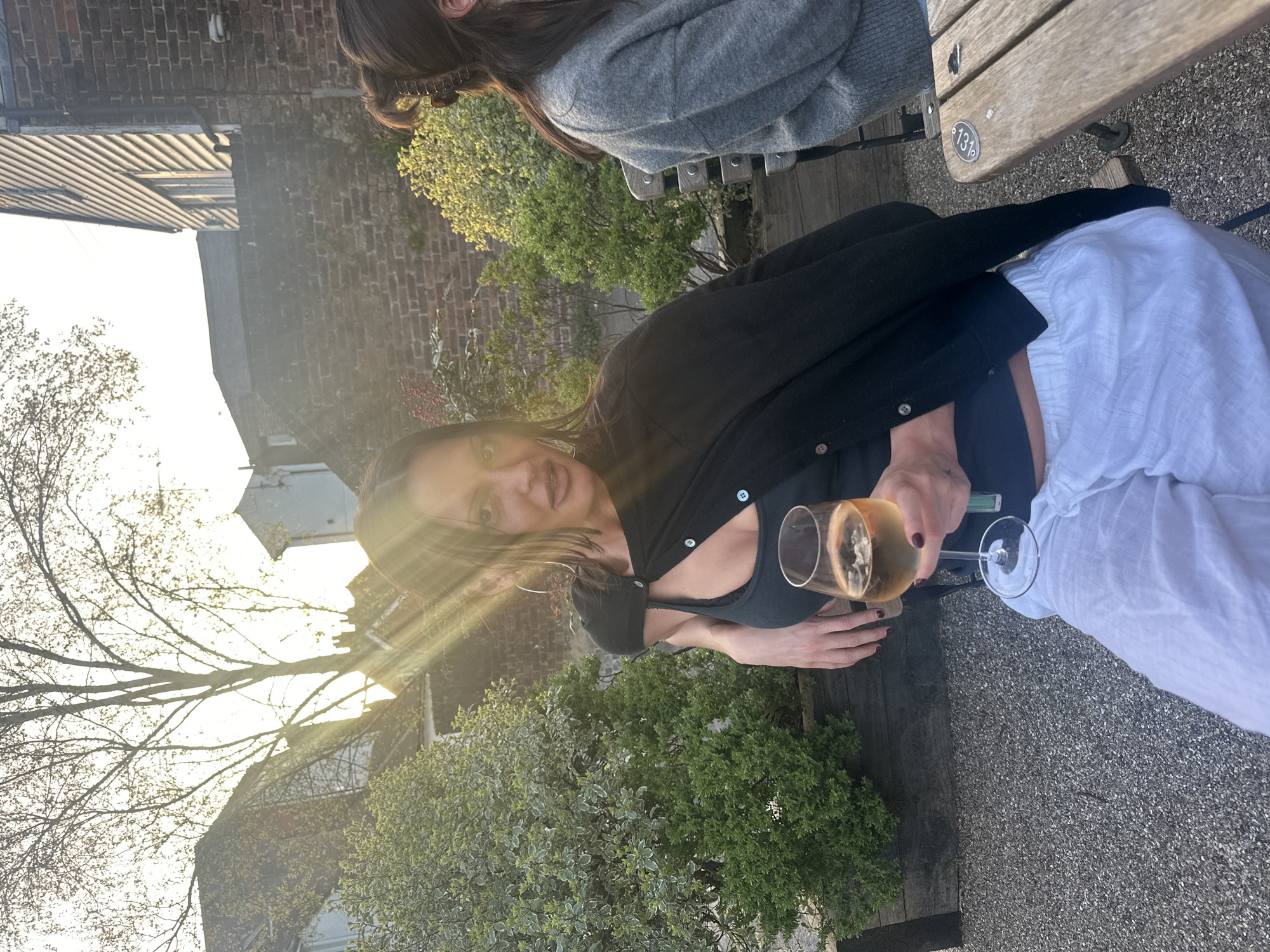 Here is a description of the image:
A woman is sitting on a bench outside, holding a glass of white wine. She is wearing a black cardigan, a blue top, and white pants. Another person is partially visible on the right side of the image. The background features foliage, buildings, and the sun shining through the trees. The image appears to be taken in Tunbridge Wells, United Kingdom.