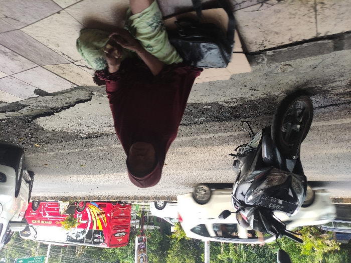 The image depicts an outdoor street scene in Situbondo, Indonesia, taken during the daytime. The primary subject is an older woman, wearing a red headscarf (hijab) and light green patterned pants, sitting on a cracked tiled sidewalk. Her face is visible, looking towards the viewer, with a somewhat neutral or somber expression. Next to her on the sidewalk is a black bag.
The woman is positioned next to a paved road with visible cracks and wear. On the road, a black scooter is parked in the foreground, facing right, with its front wheel angled. Its license plate clearly displays "P 2093 F" and "10 - 27", indicating its registration details. In the background, there is ongoing traffic, including several white cars and a distinct red public transport vehicle, likely an "angkot," which features yellow accents and text. The text on the red vehicle reads "SELAMAT DATANG DI SITUBONDO" (Welcome to Situbondo) and "GENTONG BIRU." Further in the background, trees and other buildings can be vaguely seen, suggesting an urban or semi-urban setting. The overall scene conveys a candid moment of street life.