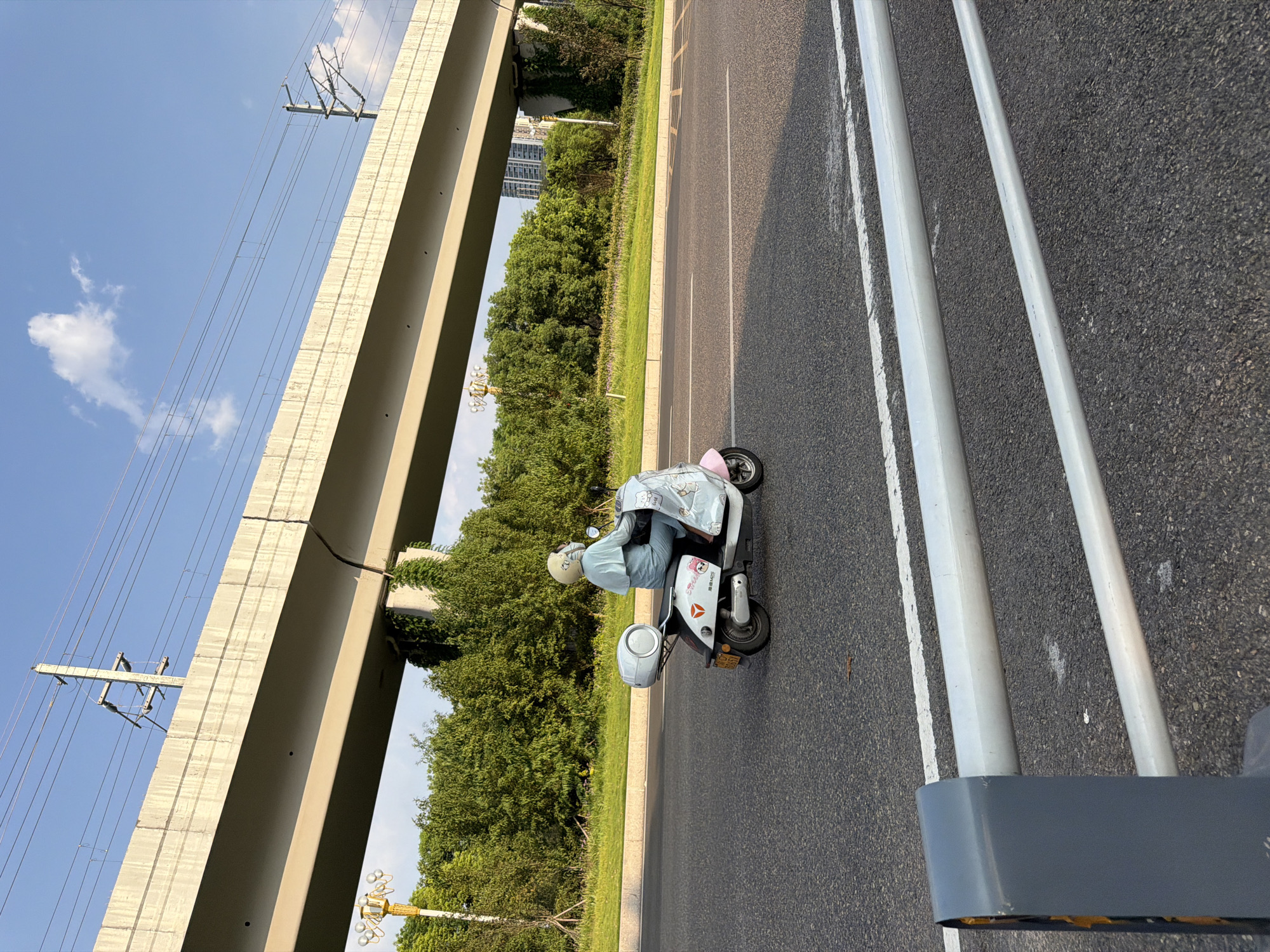 A person wearing a light blue long-sleeved shirt, light blue pants, and a white helmet is riding a white and pink electric scooter on an asphalt road. The scooter has a white storage box mounted on the back and is decorated with cartoon cat stickers. The scooter is moving from left to right in the frame. In the background, there is a large concrete overpass with electrical wires running along its top. Lush green trees and vegetation line the side of the road. Further in the background, there are some tall buildings visible. The sky is blue with a few scattered clouds. The scene appears to be a street in Jiangjin, China, possibly during the daytime given the lighting. In the foreground, there are metal guardrails.