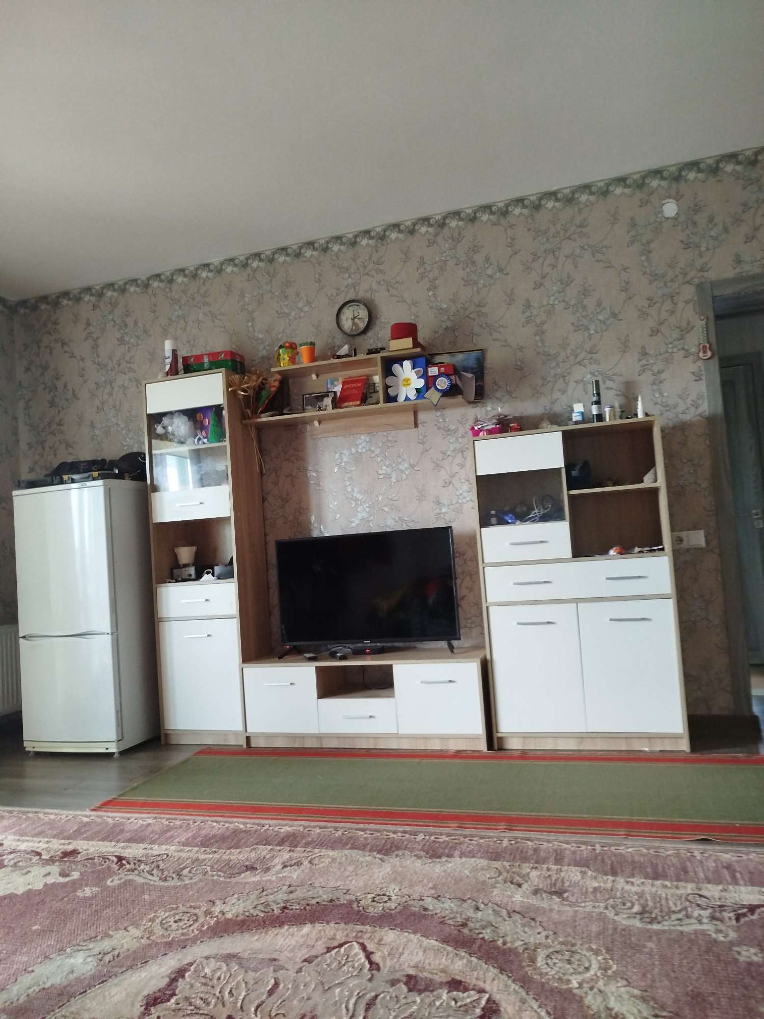The image shows a living room with a refrigerator on the left, a tall display cabinet with white doors and wooden shelves, and a television stand with white drawers and wooden accents on the right. The walls are covered with floral wallpaper. On top of the display cabinet and television stand are various objects, including boxes, toys, and bottles. A large rug covers most of the floor, with a smaller green rug placed on top of it in front of the television stand. The room appears to be brightly lit, suggesting it is daytime. The overall impression is that of a lived-in, functional living space. There is no visible text in the image.