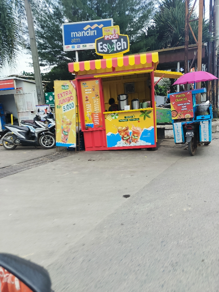 The scene is a street in Ngamprah, Indonesia, located at the coordinates -6.864 latitude and 107.51 longitude. There is a yellow and red food stand with a sign that reads "Es Teh Poci" and "Extra Jumbo Rp. 5,000." A person is standing behind the counter, presumably preparing food or drinks. To the right of the stand is a small cart with a pink umbrella on top. It is selling "Penthol Cilor 151". There are two motorcycles parked in the street. The road is made of asphalt and there is a metal grate next to the food stand. The weather appears to be sunny. There are trees in the background. The scene is typical of a busy street in a city in Indonesia.