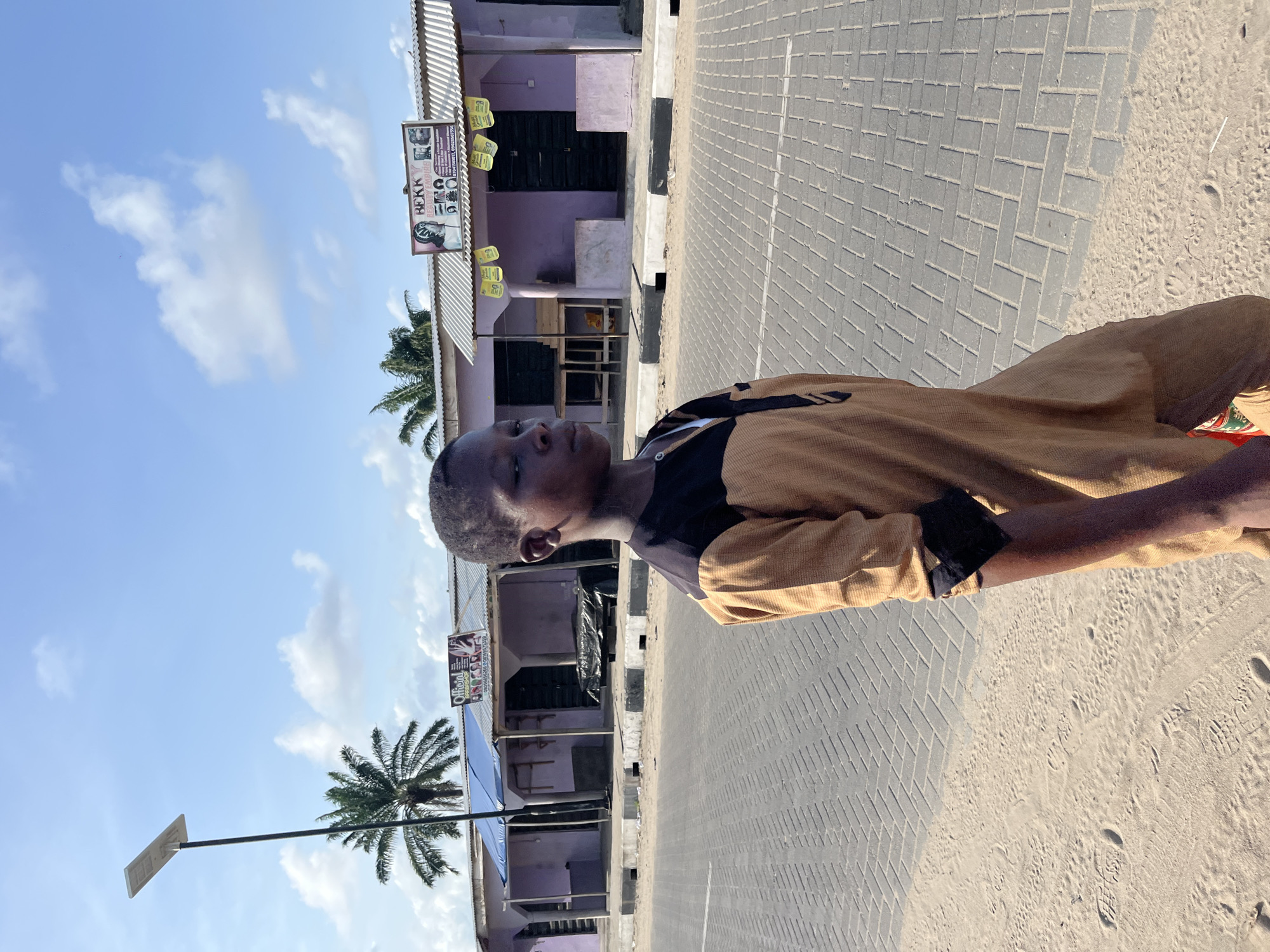 A young Black male stands outside on a bright, sunny day in Badagry, Nigeria. He is wearing a yellow and black shirt. Behind him is a paved street and a row of single-story buildings, some painted purple. There is a business sign on one of the buildings that reads "Hecky Hair Empire." Palm trees are visible in the background. A solar-powered street light is also visible. The ground on the right is sandy.