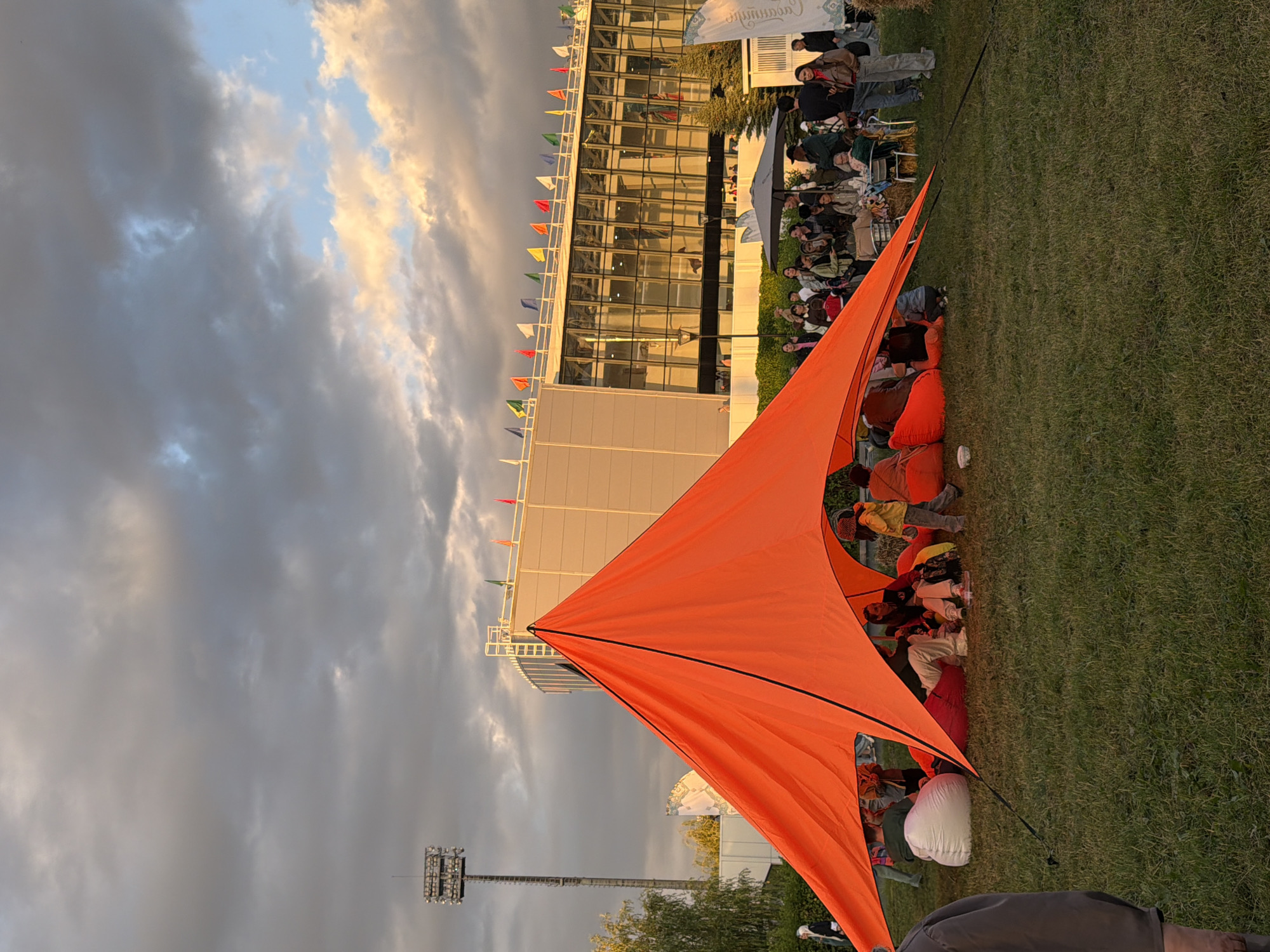 An outdoor gathering is taking place in front of a modern building with a glass facade and a row of flags along its roofline. In the foreground, an orange tarp is pitched, creating a shelter. Beneath the tarp and on the surrounding lawn, several people are seated on beanbag chairs, some engaged in conversation. The scene is illuminated by warm, late afternoon or early evening light, casting long shadows and highlighting the golden hues of the sky. A tall floodlight tower stands to the left of the frame, and trees are visible behind the building. The ground is covered in green grass.