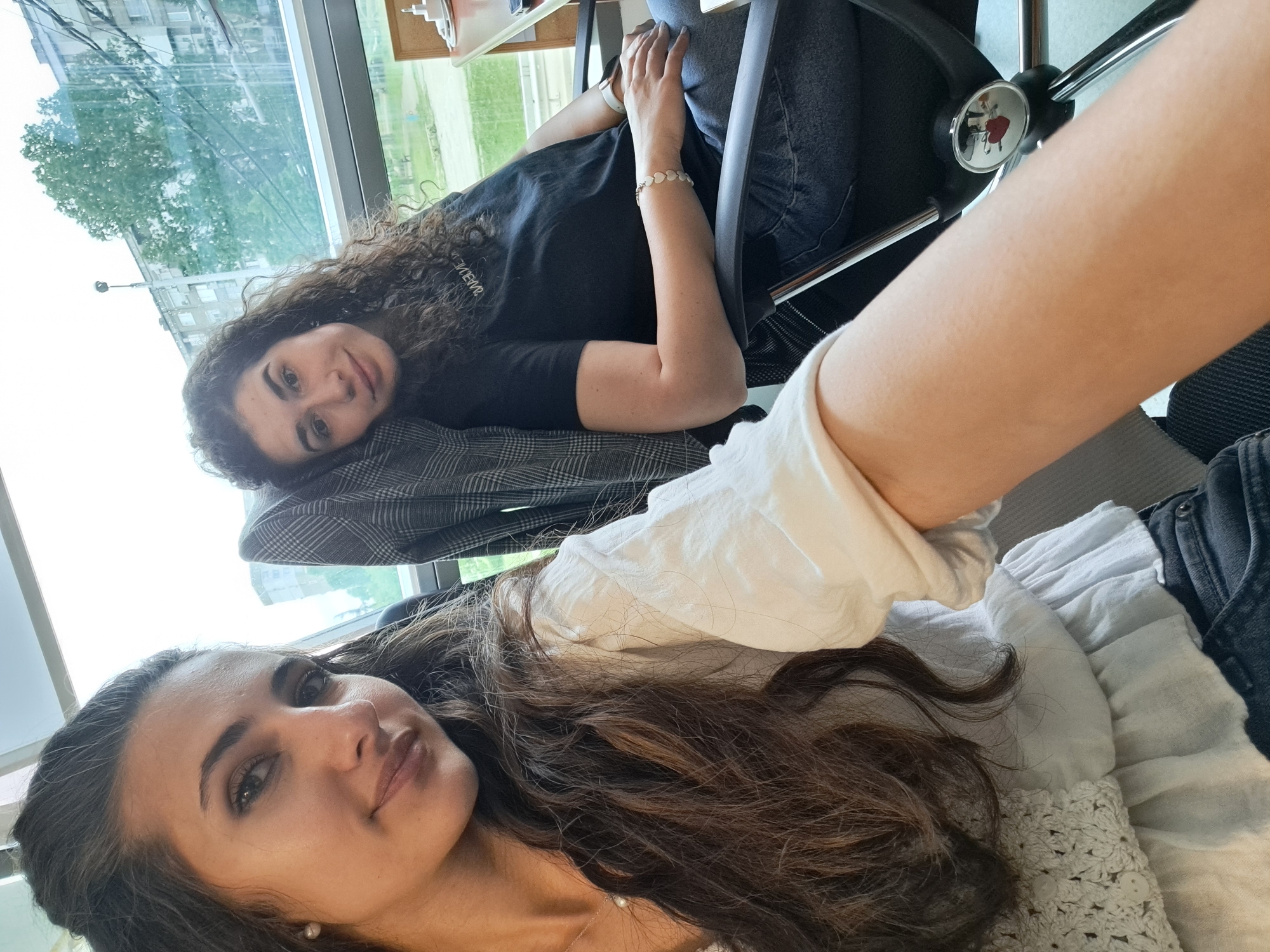 Two women are lying down indoors. The woman on the top, with curly dark hair, is wearing a black t-shirt and blue jeans. She is resting her head on a grey plaid office chair. The woman below has long brown hair and is wearing a white shirt. The location appears to be an office with large windows, possibly in Sofia, Bulgaria, based on visual clues. Outside the window, there are buildings and trees.