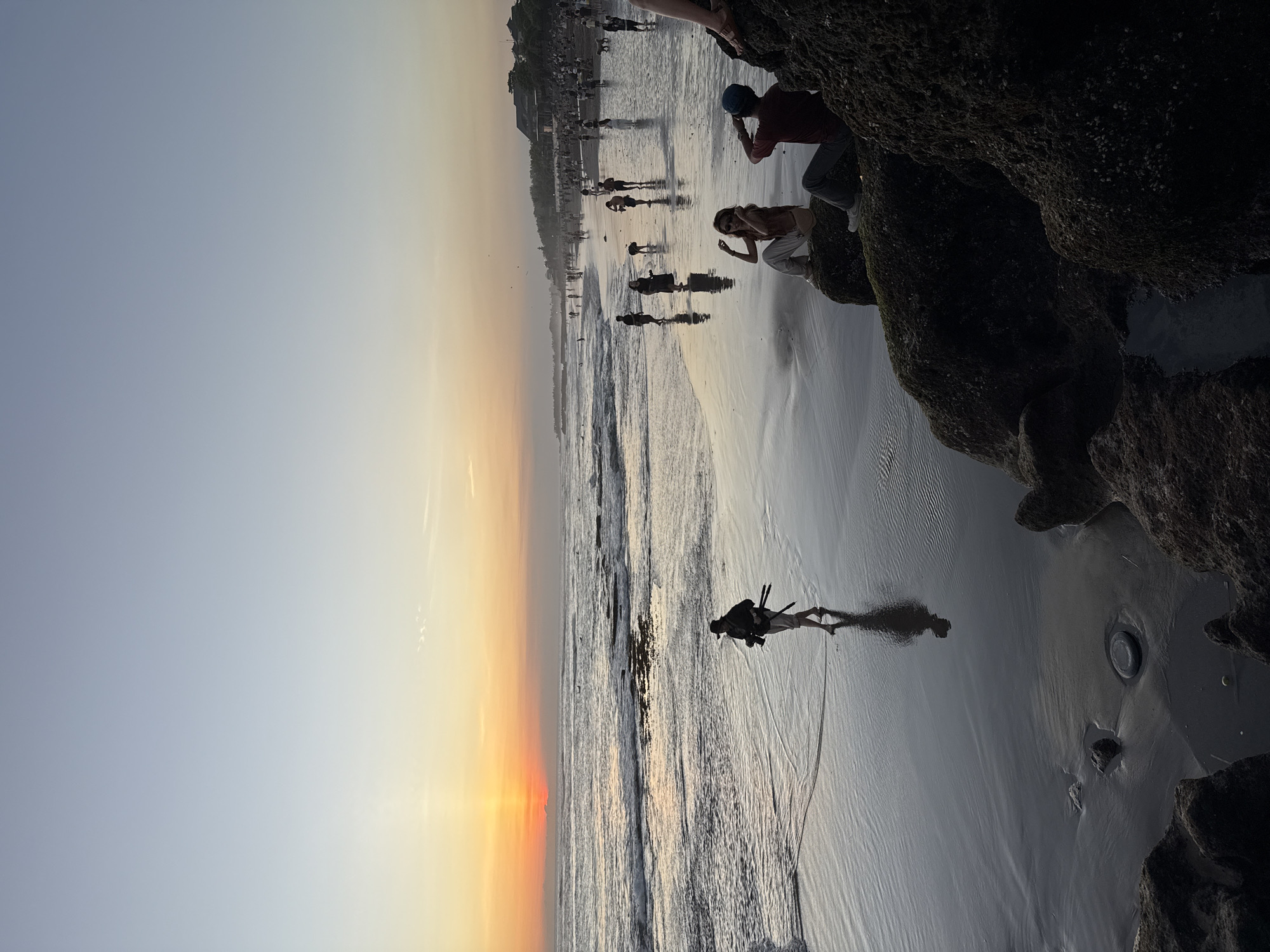 The image captures a serene sunset scene at a beach in North Kuta, Indonesia. The sun is setting on the left side of the frame, casting warm orange and yellow hues across the horizon and reflecting on the wet sand and gentle waves of the ocean. The sky transitions from vibrant sunset colors to a softer blue-grey overhead.
Numerous people are scattered across the wide sandy beach, enjoying the evening. Several individuals are visible closer to the camera, including a person in the shallow water on the left, holding a camera mounted on a tripod, silhouetted against the setting sun. On the right side of the frame, large, dark, textured rock formations jut out from the sand. Two people are perched on these rocks; one person in a red shirt and blue cap appears to be looking out at the ocean or taking a photo, while another person in light-colored clothing is also situated on the rocks. Further down the beach, more people can be seen walking, wading, or standing near the water's edge, their figures forming various silhouettes against the diminishing light.
The scene conveys a peaceful atmosphere, indicative of leisure activity during the golden hour. The lighting creates a strong contrast between the bright sky and the darker foreground elements, highlighting the silhouettes of the people and rocks. There is no visible text in the image.