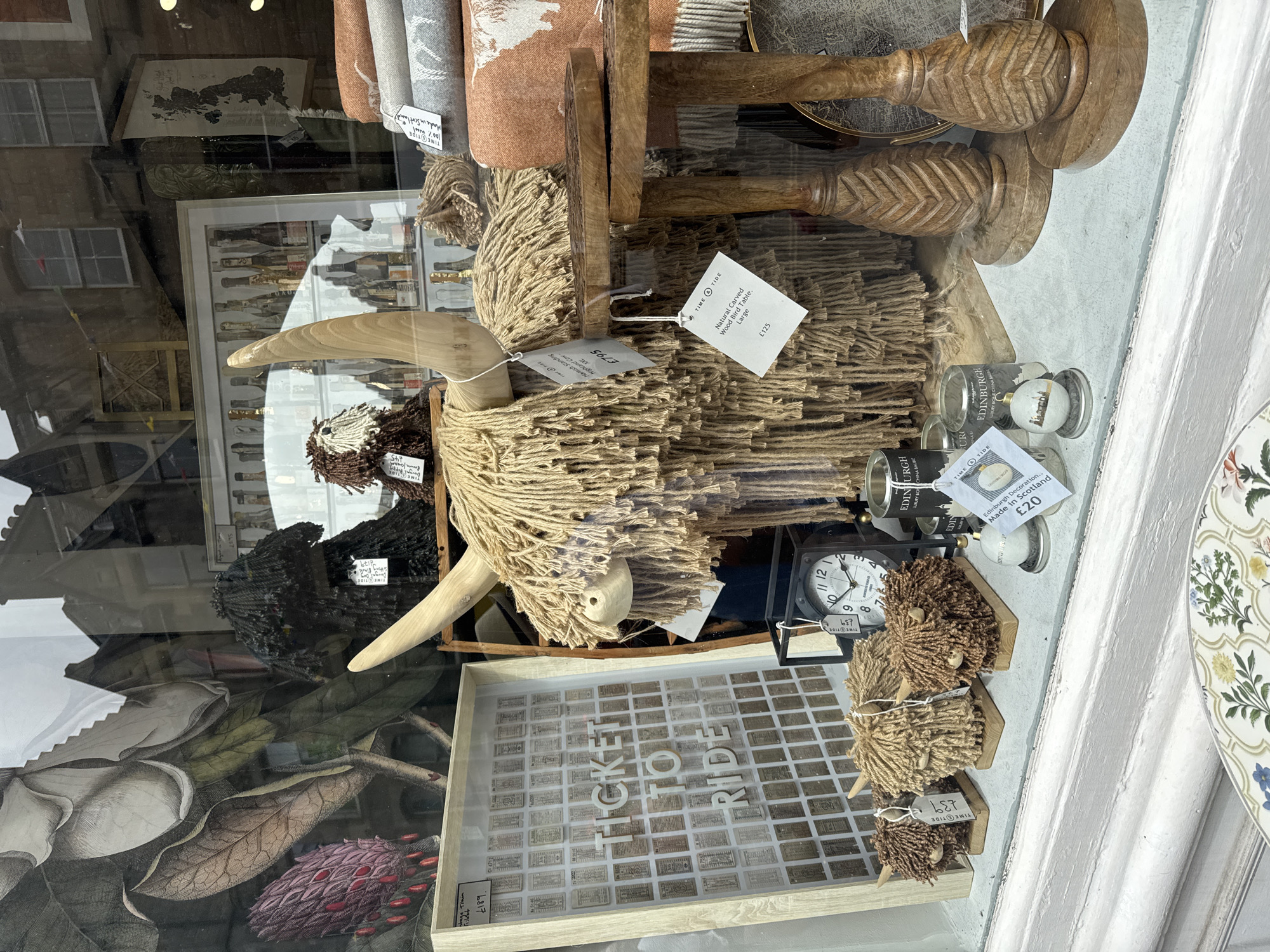 This is a close-up, eye-level, outdoor photograph of a shop window display in the City of Edinburgh, United Kingdom. The display features several decorative items, primarily focusing on Highland coo (cow) figures and textiles.
In the foreground, to the left, is a framed display containing a grid of vintage-looking tickets, with the words "TICKET TO RIDE" in raised white lettering. To the right of this is a large, shaggy Highland coo figure made of natural fibers with prominent wooden horns. Several smaller, similar Highland coo figures are positioned in front of it. A dark-faced clock with a white face and Roman numerals is visible behind the smaller coo figures. Various cylindrical containers, possibly candles or diffusers, are also present, some with labels indicating "EDINBURGH".
Further back, on the right side of the frame, are stacked textiles, including what appear to be blankets or throws in shades of orange and grey, some with white patterns. Two carved wooden side tables with intricately patterned bases and tops are also part of the display. A large botanical illustration featuring a purple flower and large leaves is visible on the far left, behind the ticket display. Reflections of buildings and the sky are visible in the shop window glass.