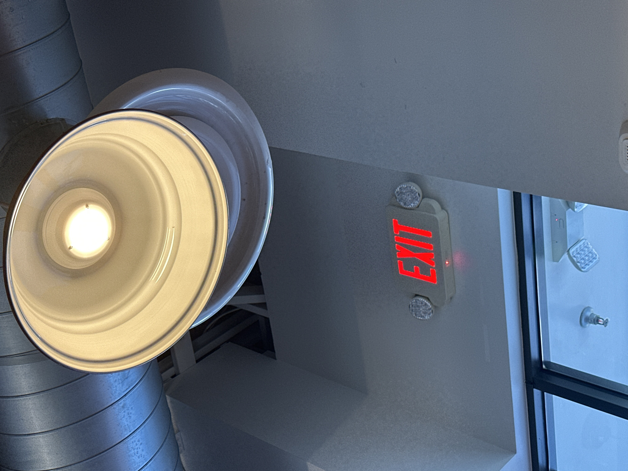 This image shows an interior view of a ceiling with a large industrial-style light fixture and a red illuminated "EXIT" sign. The light fixture is a wide, conical shade with a bright bulb at its center, casting a warm glow. Below the light, a white rectangular exit sign with red lettering is mounted on the ceiling. The sign also has two small, circular emergency lights on either side. In the background, a large, corrugated metal duct runs horizontally. Below the exit sign, a portion of a large window or glass partition is visible, reflecting a bright blue light. A small fire sprinkler head is also visible mounted on the ceiling in the reflected light. The overall environment appears to be an indoor public space, possibly a commercial building or industrial facility. The image was taken in Lynnwood, United States of America.