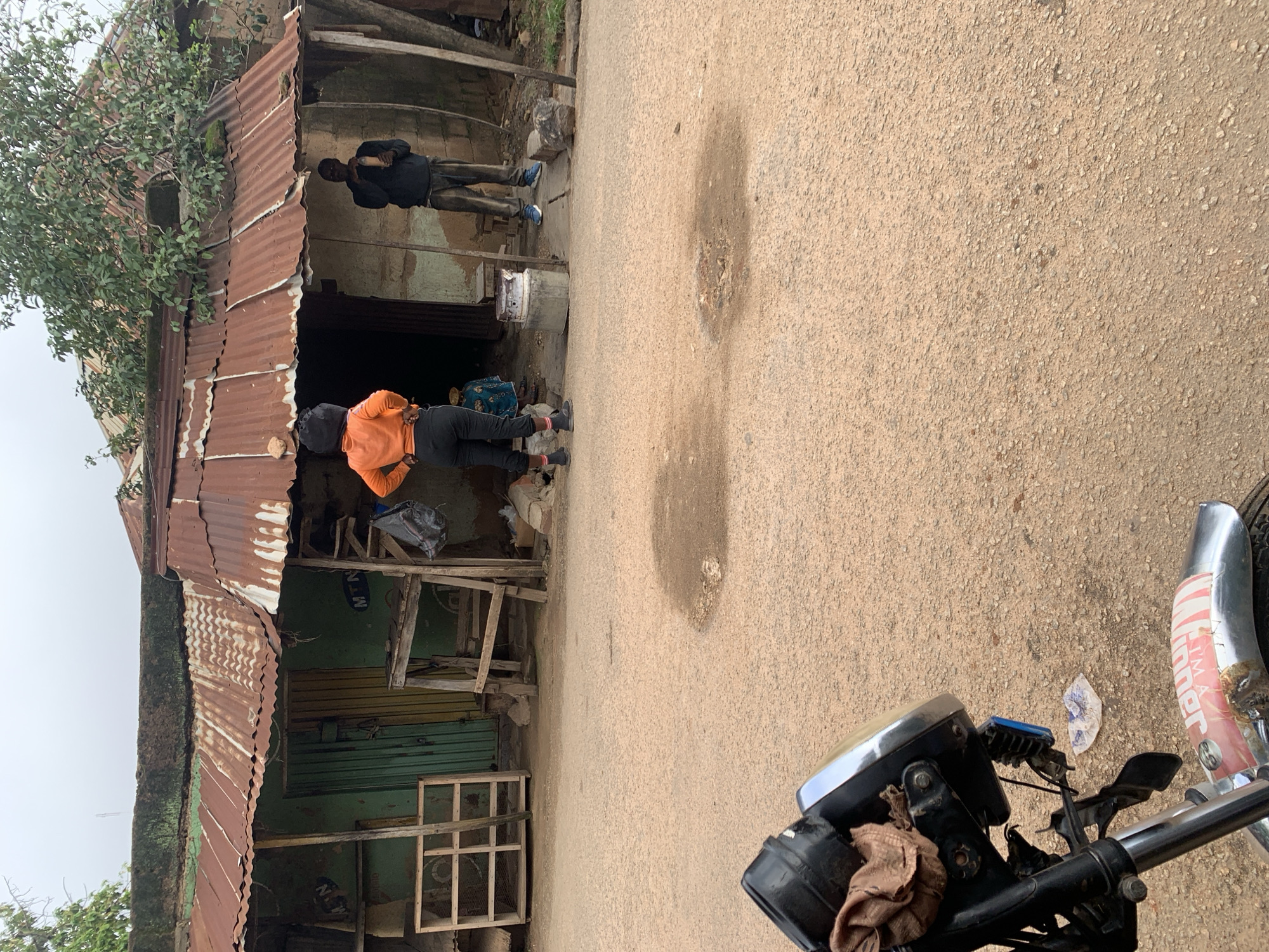 The image shows a street scene in a rural or semi-urban area. In the foreground, the front of a motorcycle is visible, with its headlight and handlebars taking up the lower left portion of the frame. A brown cloth is tied to the headlight. The motorcycle is parked on a dirt or gravel road, which is somewhat uneven and has a few puddles or damp patches.
In the middle ground, there is a row of structures that appear to be shops or market stalls. These structures have corrugated metal roofs that are rusty and weathered. The walls are made of a mixture of materials, including what looks like mud or concrete blocks. One stall has a green door and a yellow door. Another stall has a wooden frame for displaying goods.
Two individuals are standing in front of the stalls. One person, wearing an orange hooded jacket and dark pants, is standing with their back to the camera, facing the stalls. They have their hands on their hips. The second person, wearing a dark hooded sweatshirt and grey pants, is standing to the right, facing the stalls and holding a light-colored object. A third, smaller figure in a blue and white patterned garment is partially visible behind the person in orange.
In the background, there is vegetation, including a tree with green leaves growing over one of the structures. The sky is overcast, suggesting a cloudy day. The overall impression is one of a simple, everyday street scene in a location with basic infrastructure.
