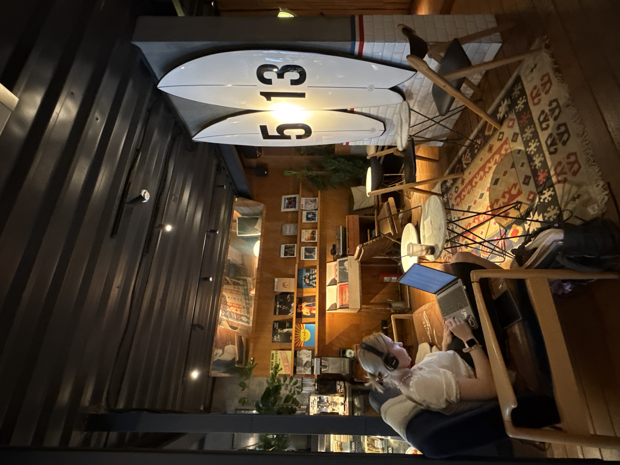 This image captures a cozy and eclectic interior space, possibly a cafe or a creative lounge, featuring a blend of industrial and beach-inspired decor. The scene is warmly lit, suggesting evening or low light conditions.
The primary subject is a person with light-colored hair, wearing headphones and a light top, seated comfortably in a wooden armchair or sofa. They are actively engaged with a silver laptop, typing, indicating work or leisure activity. A smartwatch is visible on their left wrist.
The background features a prominent dark corrugated metal wall on the left, contrasting with a wooden panel ceiling and white tiled sections. Two white surfboards are mounted horizontally on a wall or beam above the tiled area, each bearing the black numbers "513" on their underside. A patterned rug in reds, blacks, and creams covers part of the floor.
To the person's right, a wooden shelving unit is filled with numerous record albums and books. Visible album titles include "MORCHEEBA," "THE CURE," "SEX PISTOLS," "NEVER MIND THE BOLLOCKS HERE'S THE SEX PISTOLS," and partially "MILES DAVIS" and "THE RAMONES." A plant is also visible on a lower shelf. The overall ambiance is relaxed and inviting, a conducive environment for focused work or enjoyment, set within what could be a unique establishment in Moscow, Russia.