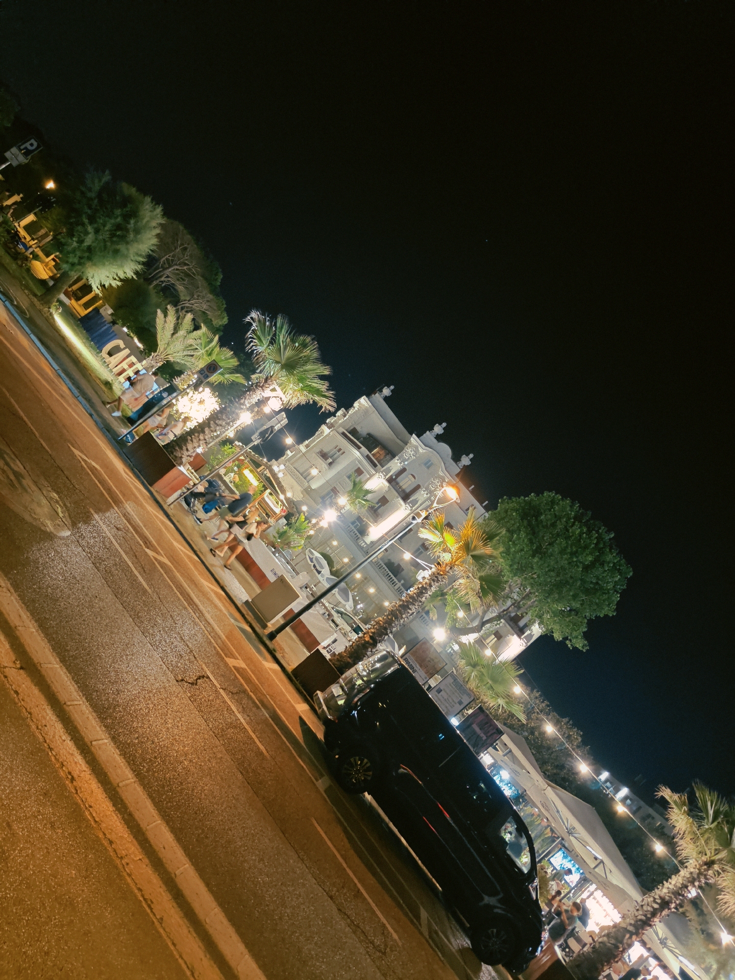 This is a night time, eye-level, oblique shot of a street scene in Rimini, Italy. The shot captures a building with several floors, featuring decorative architectural elements and illuminated windows. In front of the building, there is a cafe or restaurant area with outdoor seating, palm trees, and string lights. A black car is parked on the right side of the street. The street itself is paved and illuminated by streetlights, casting a warm glow on the wet surface. Several people are visible, some walking, others seated at tables, appearing to be patrons of the establishment. A large sign with the word "CAFE" in white block letters is visible on the left side of the frame. The sky is dark and clear, indicating it is nighttime.