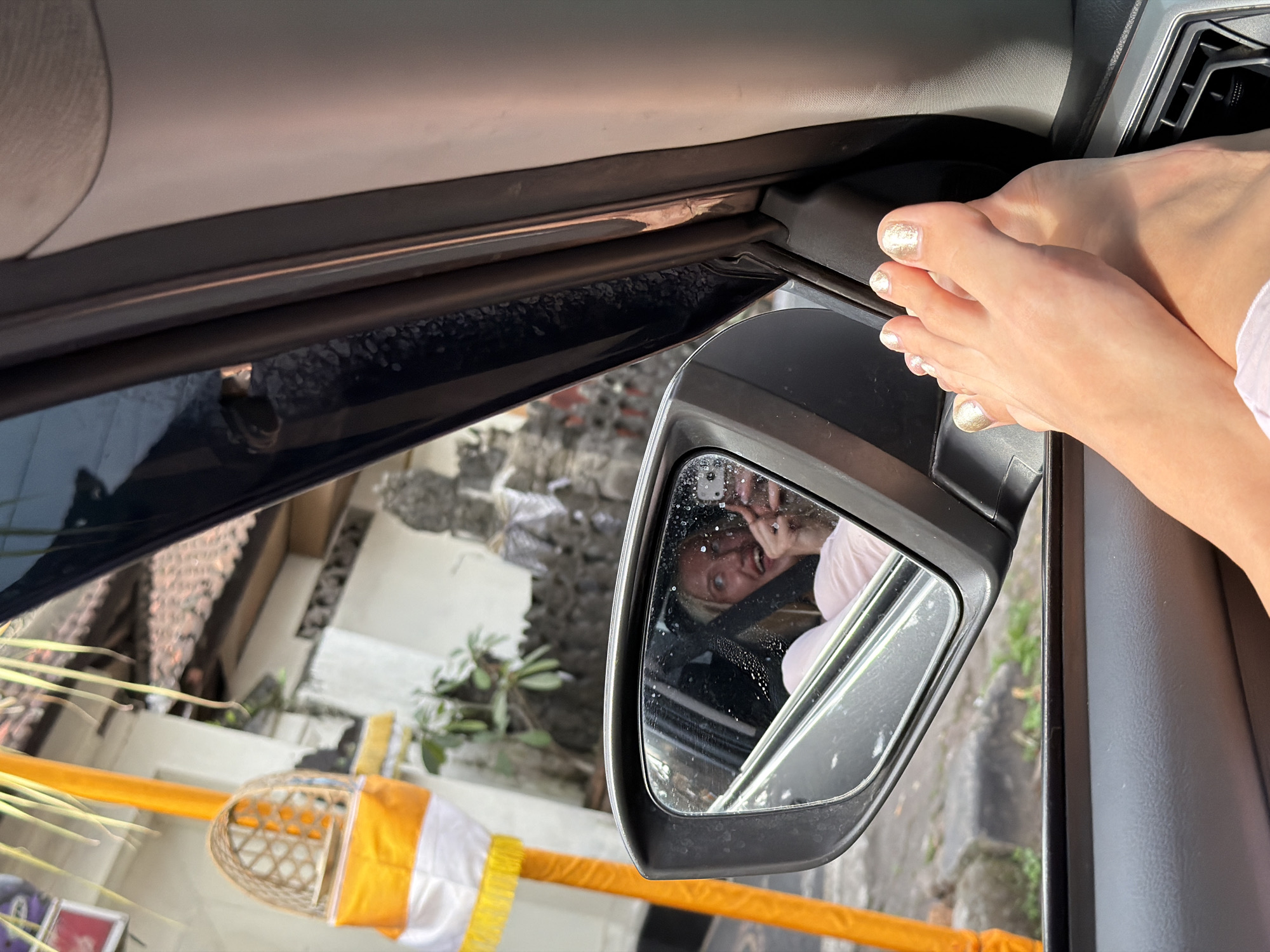 In the city of Mengwi, Indonesia, a woman is inside a vehicle, her bare feet resting on the dashboard. She is looking into the side-view mirror, which reflects her face with a surprised expression. She appears to be taking a selfie with her phone. Outside the vehicle, a traditional Balinese umbrella is visible.
