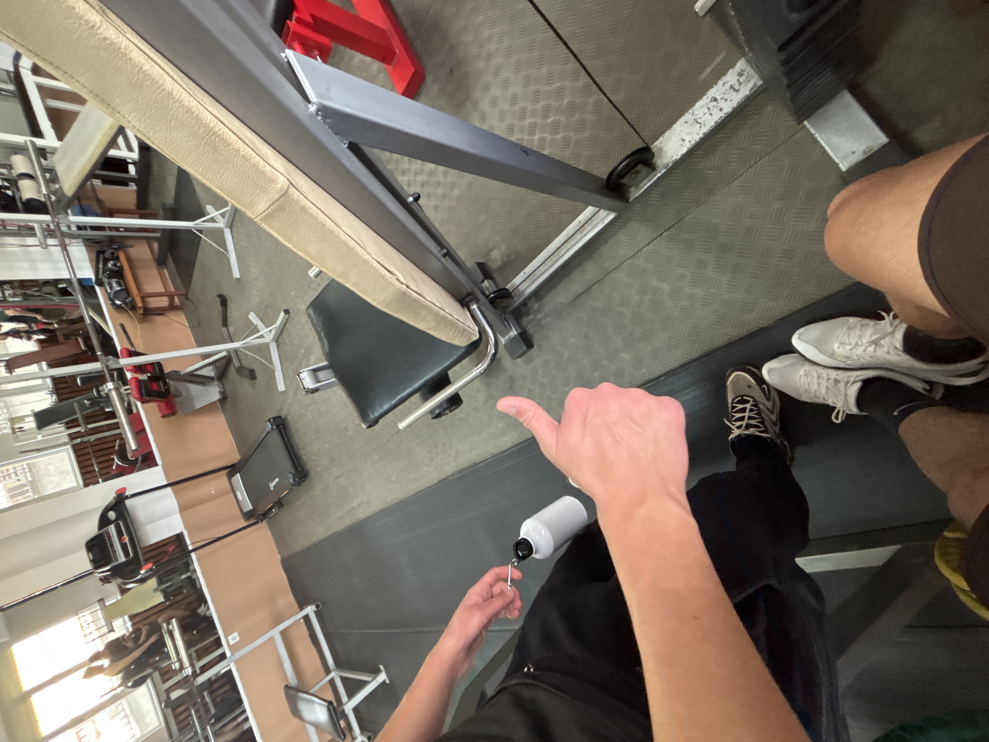 The image shows an interior gym scene. The view is a first-person perspective looking down towards the body of the person holding the camera. The person is holding a white water bottle in the left hand, giving a thumbs up with the right hand. The person is wearing black athletic pants and two different colored running shoes. To the left of the person are a workout bench, weight machines, and other gym equipment. In the background is a window that provides light to the space. The location is in Zlatograd, Bulgaria.