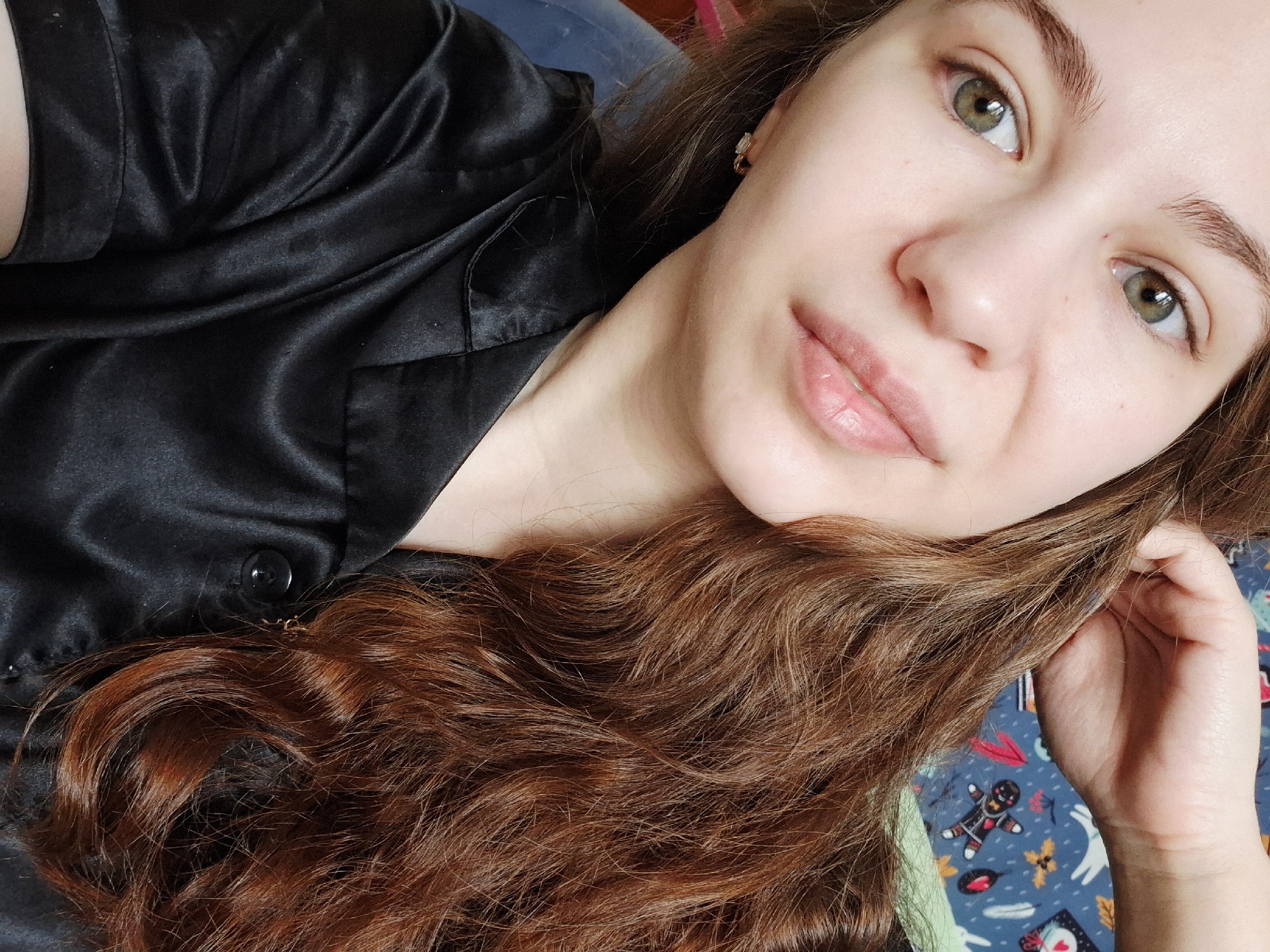 A selfie shows a woman with fair skin and hazel eyes. She has light brown eyebrows and full, glossy lips. She is wearing a black satin shirt with a button closure at the chest. Her long, wavy auburn hair is spread out around her head, and a small earring is visible in her ear. She appears to be lying down on a surface with a blue patterned fabric. The location is unspecified, and there is no discernible evidence to suggest that the image was taken in the city of Chaikovsky, Russia.