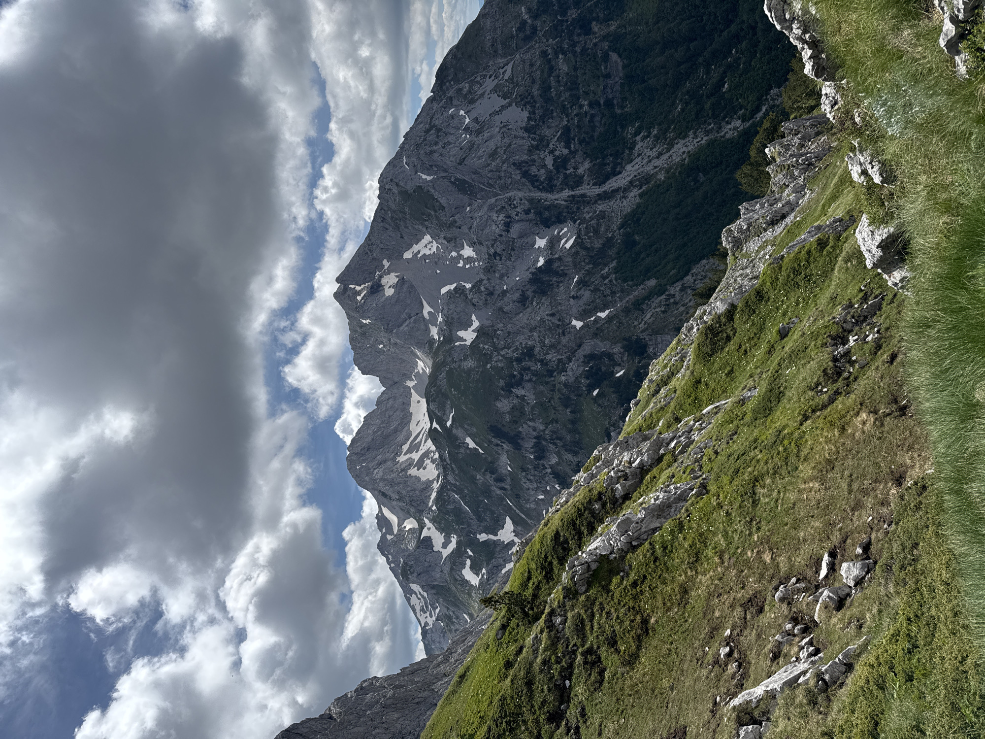 The image shows a dramatic mountain landscape in Montenegro under a cloudy sky. The foreground features a steep, grassy slope dotted with rocks. In the mid-ground, the slope continues upwards, leading to rugged, rocky peaks. Patches of snow are visible on the higher elevations of the mountains. The background consists of more distant, jagged mountain ranges, with dark green forests clinging to their lower slopes. The sky is filled with a mixture of bright white and darker grey clouds, suggesting variable weather conditions. There are no people or man-made structures visible in the image.